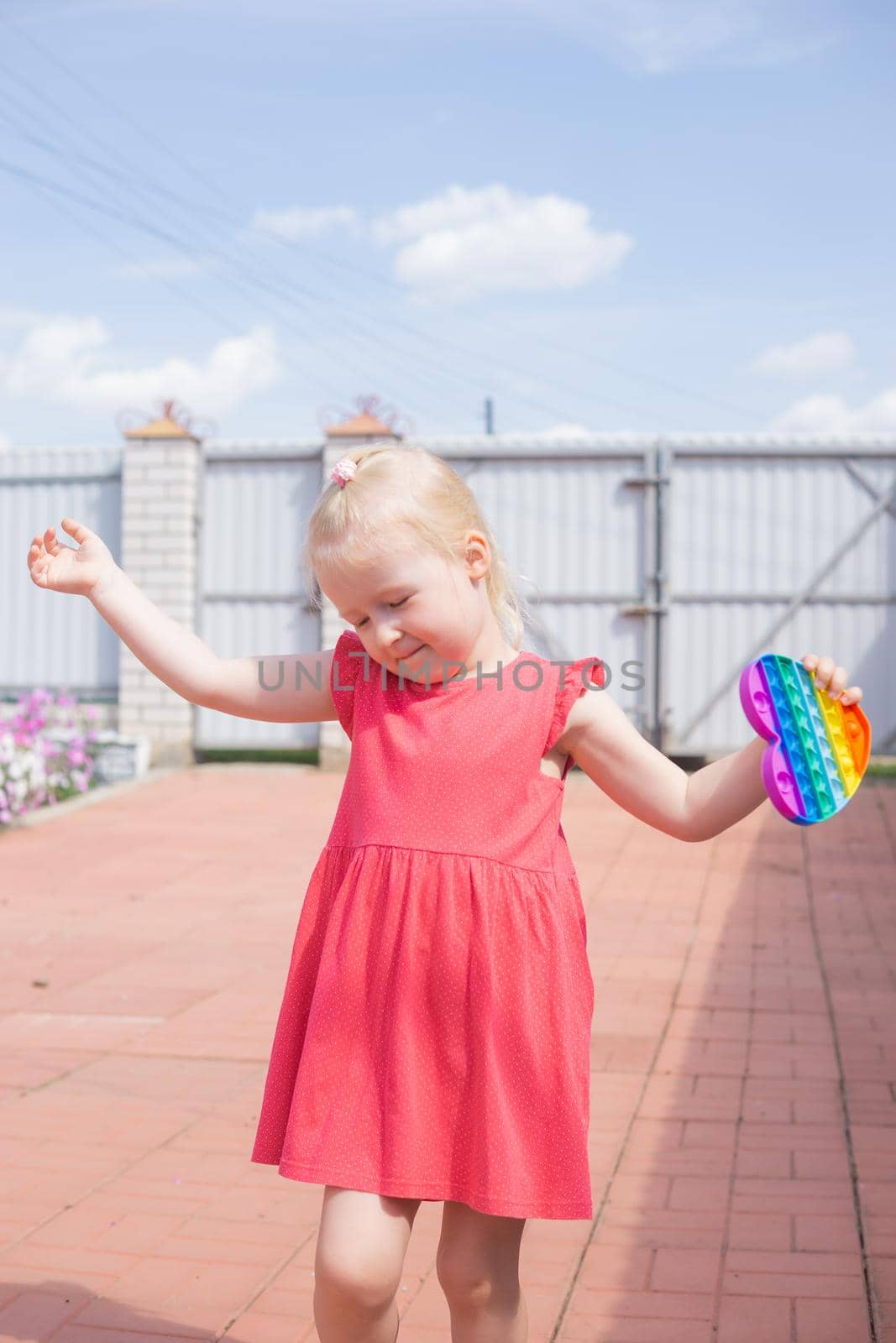 Cute caucasian fair hair girl in pink dress is playing and showing funny trendy silicone antistress colorful toy popit. Push Popper fidget toy is the ideal toy to get rid of your frustration or stress.