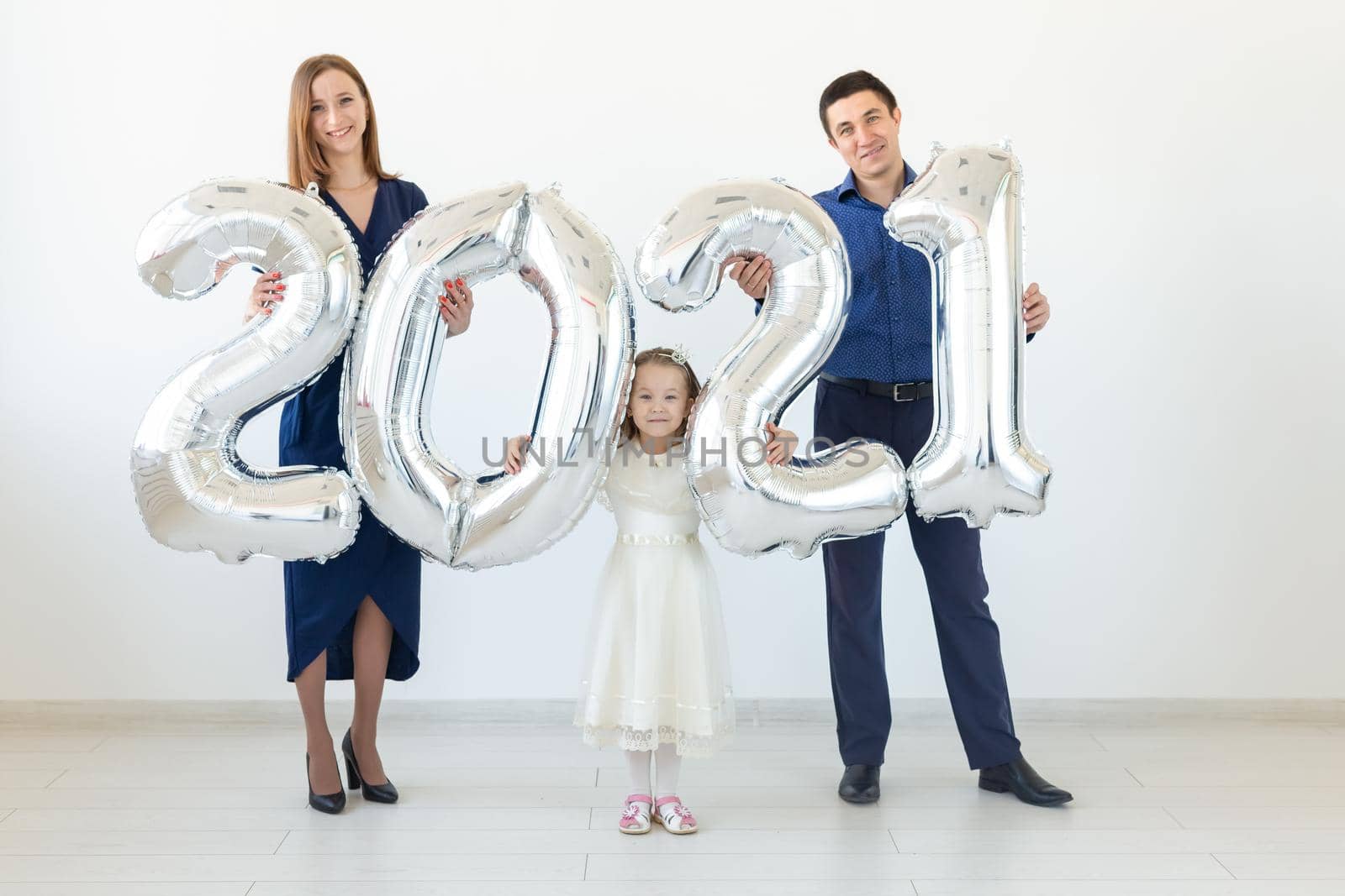 Young happy family mother and father and daughter standing near balloons shaped like numbers 2021 on white background. New year, Christmas and holiday.