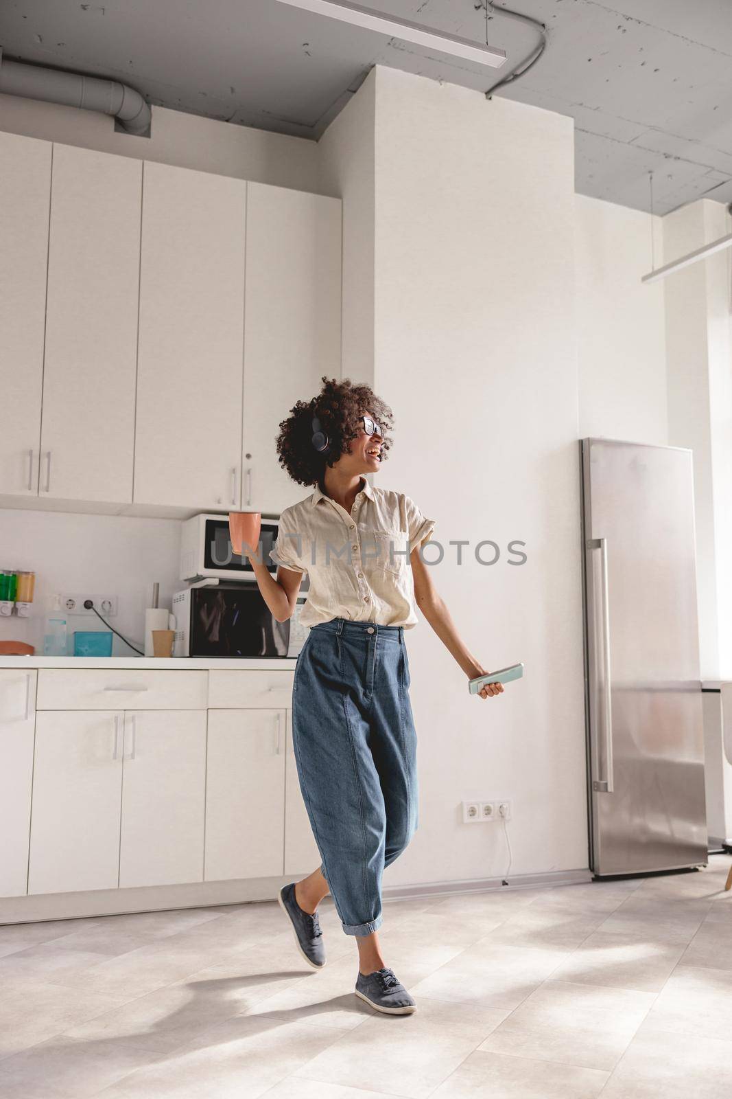 Full-length photo of smiling lady using headphones and dancing on the kitchen while holding smartphone