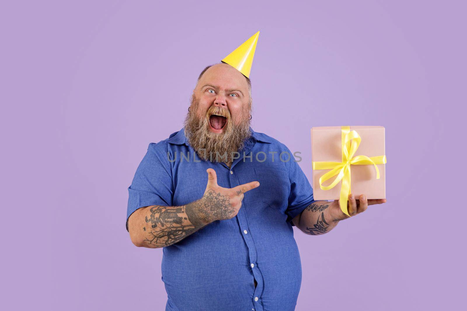 Excited middle aged man with overweight in tight blue shirt and yellow hat points onto gift box standing on purple background in studio