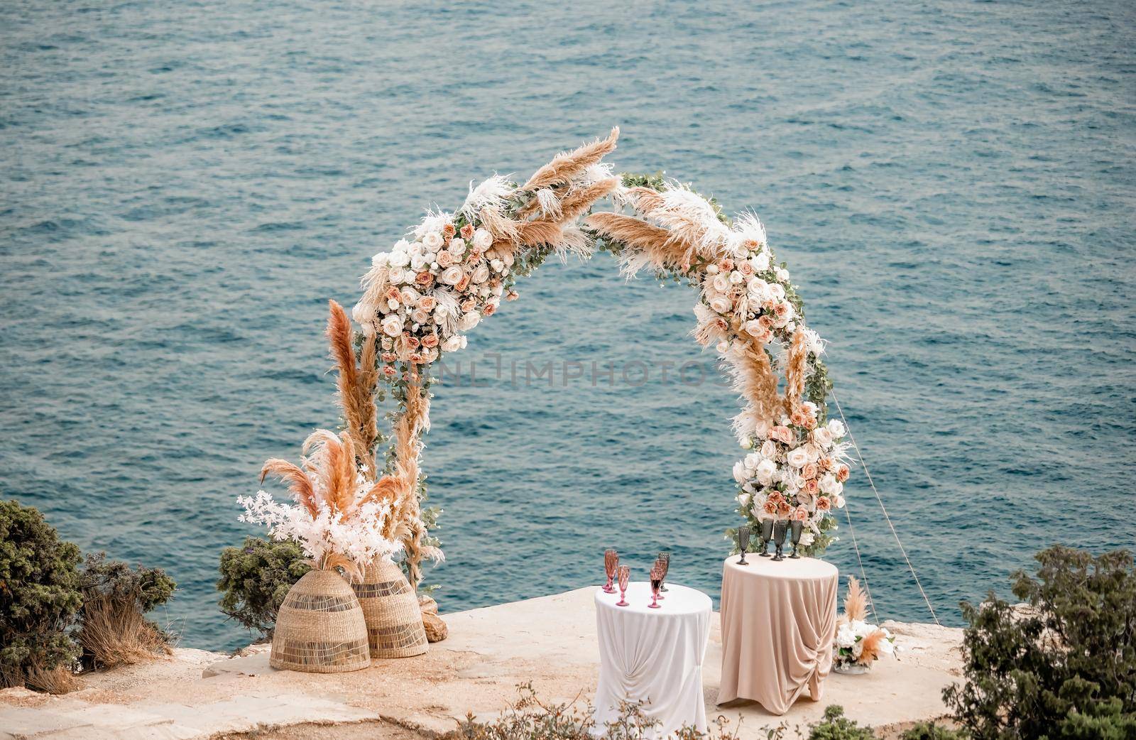The round wedding arch is decorated with a variety of flowers for beach ceremony against the sea landscape. Wedding ceremony in boho rustic style. Decor for wedding ceremony on the sea. by panophotograph