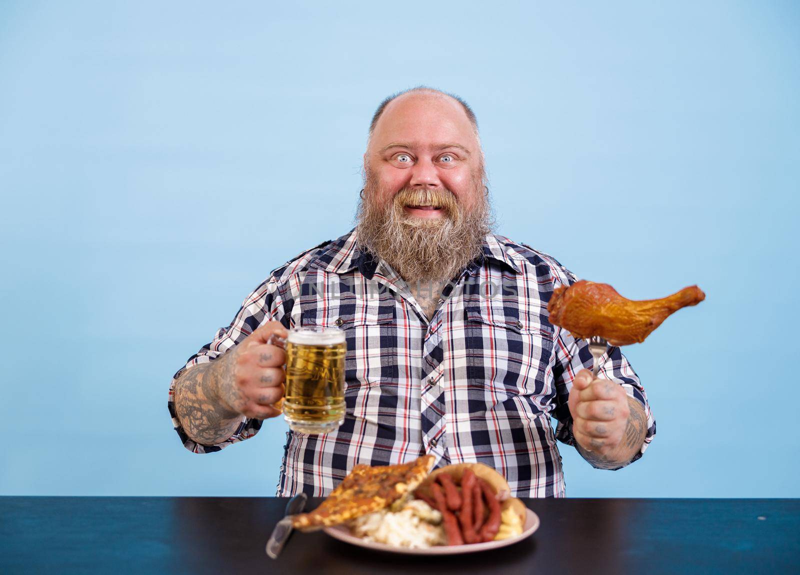 Happy plump man holds chicken leg and beer sitting at table on light blue background by Yaroslav_astakhov