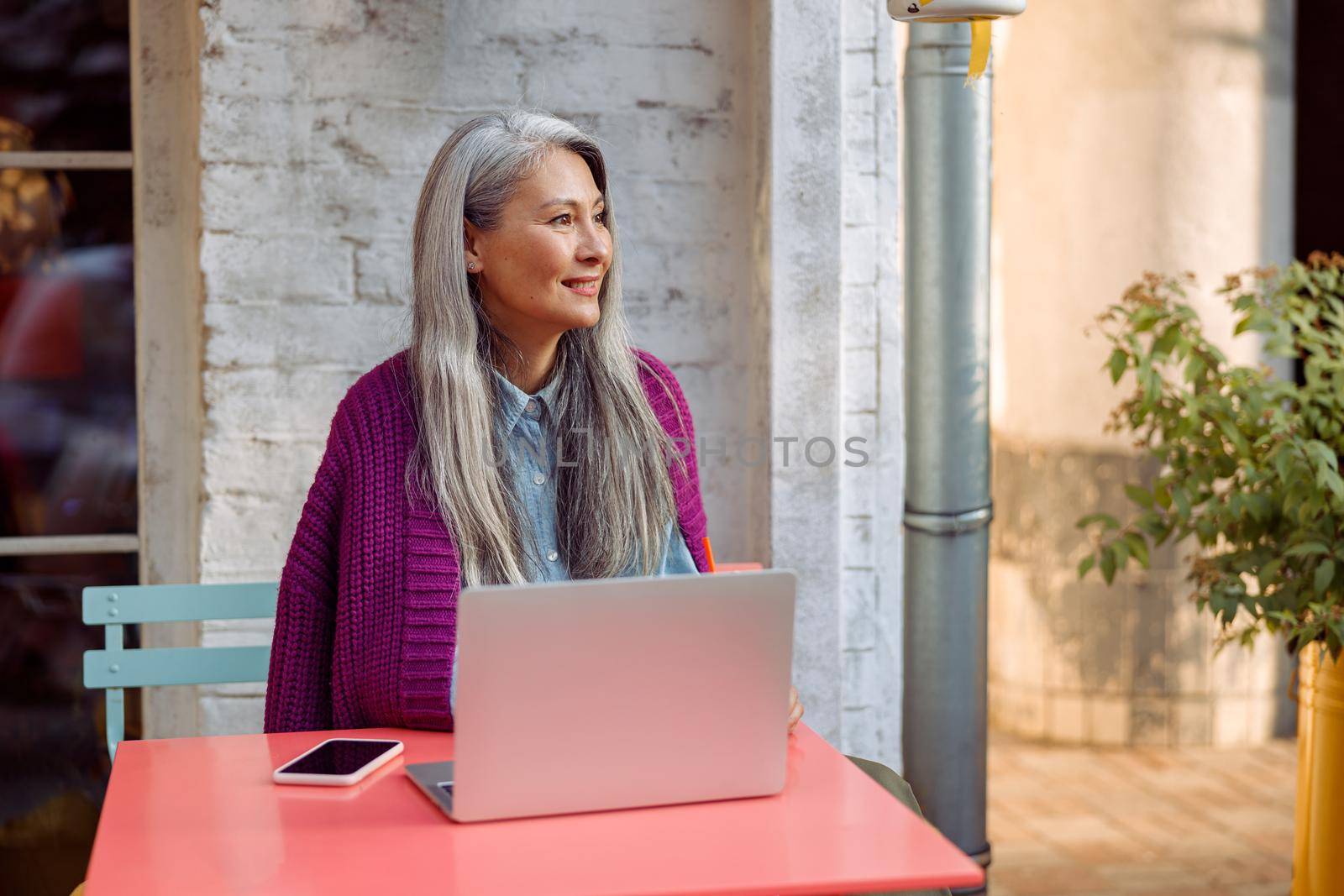 Attractive senior Asian lady in knitted jacket with smartphone and laptop sits at coral table on outdoors cafe terrace