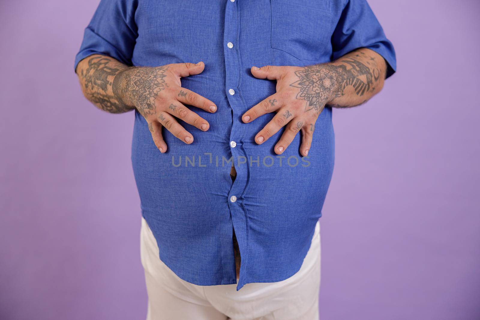 Man with overweight and tattoos on arms in tight blue shirt holds hands on large belly standing on purple background in studio closeup