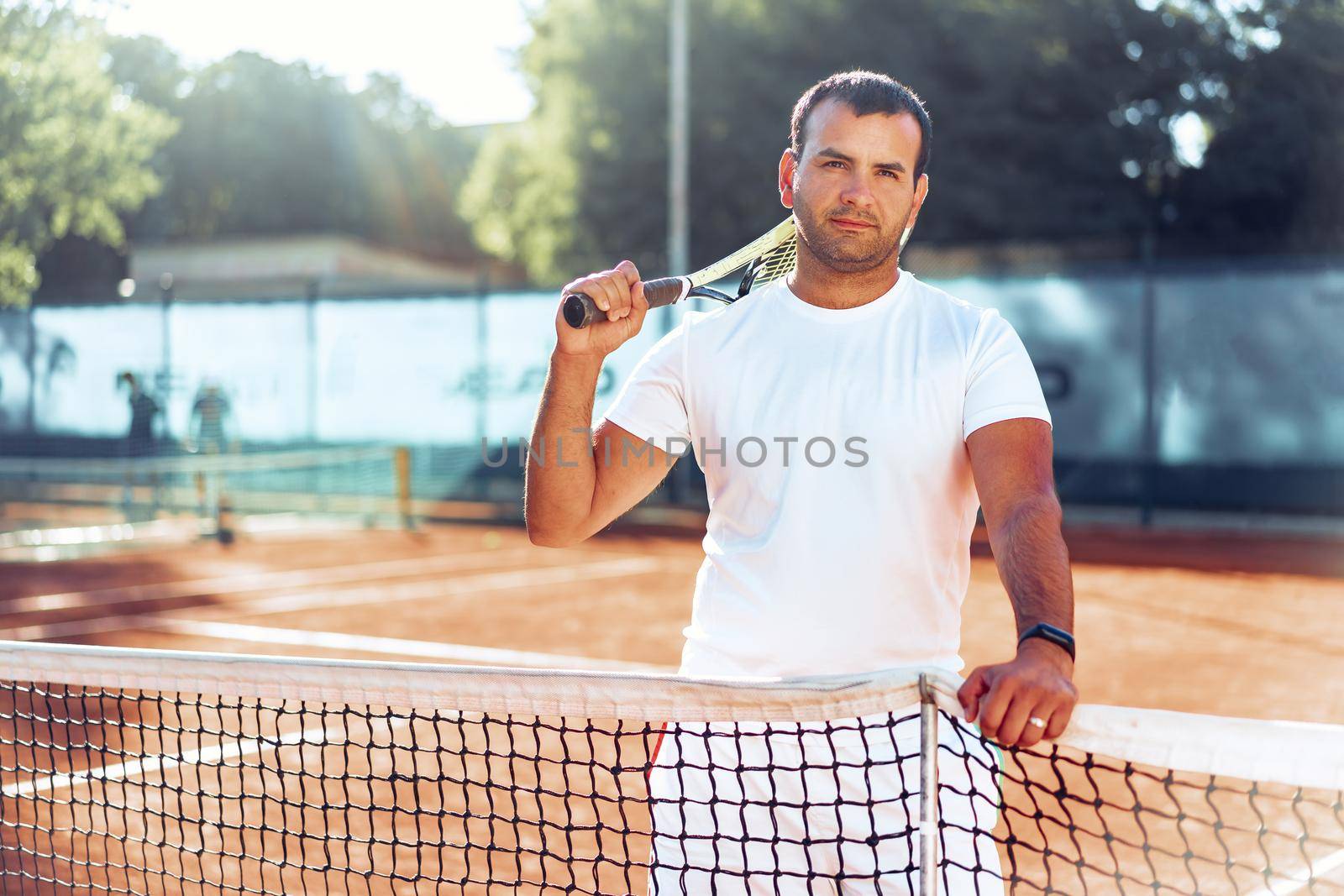 Spoty man with tennis racket standing on clay tennis court near net