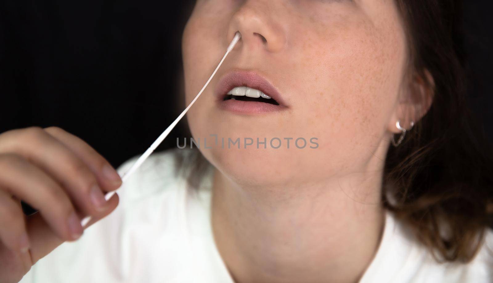 Young woman taking a Covid-19 SARS-CoV-2 test during Corona pandemic on dark black background, quick home test coronavirus closeup nose swab