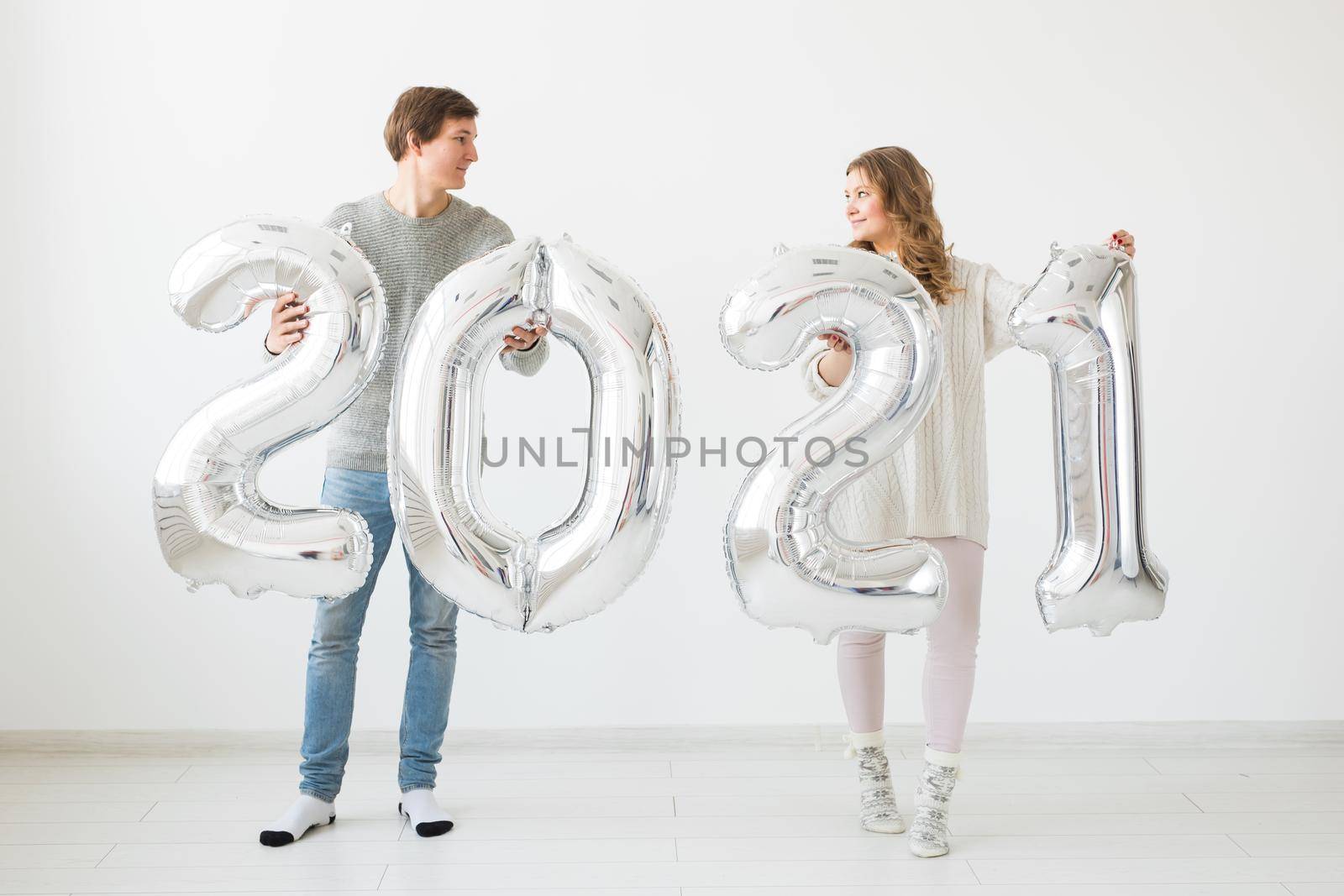Holidays, festive and party concept - Happy loving couple holds silver 2021 balloons on white background. New Year celebration. by Satura86