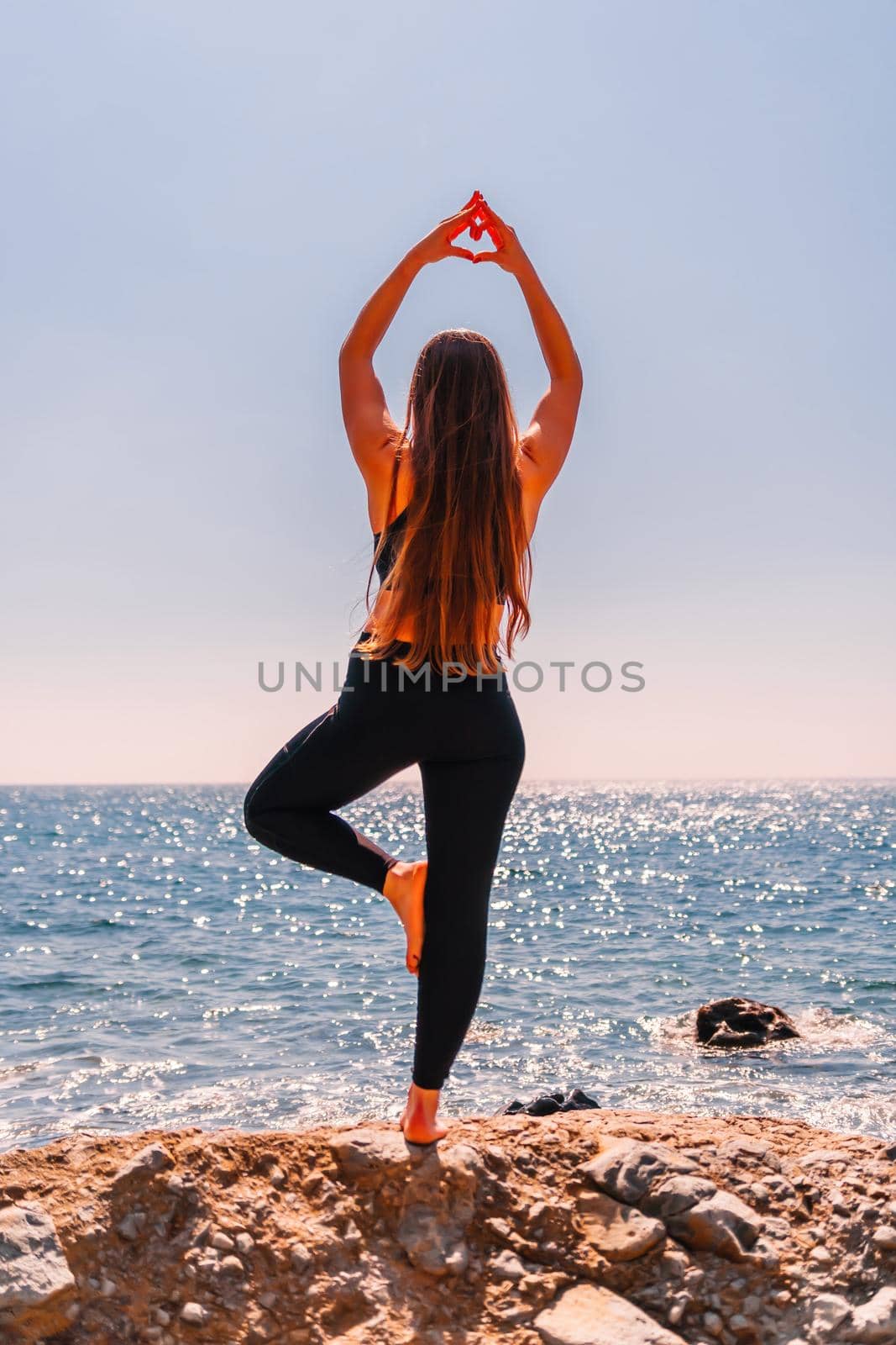 Woman makes heart with hands on beach. Young woman with long hair, fitness instructor, stretching before pilates, on a yoga mat near the sea on a sunny day, female fitness yoga routine concept by panophotograph