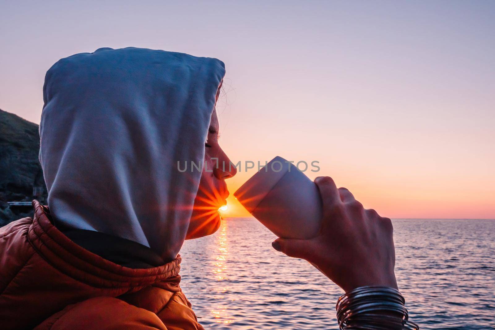 A young tourist Woman holding coffee tumbler cup while sitting outdoor and enjoying sunrise over sea mountain landscape. Women's yoga fitness routine. Healthy lifestyle, harmony and meditation by panophotograph