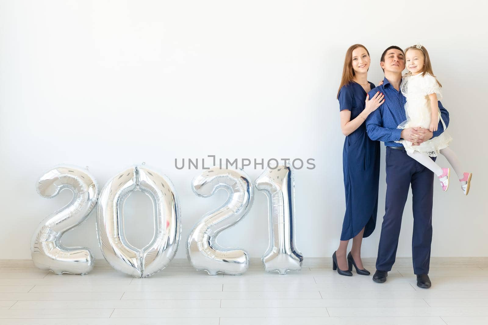 Young happy family mother and father and daughter standing near balloons shaped like numbers 2021 on white background. New year, Christmas and holiday.