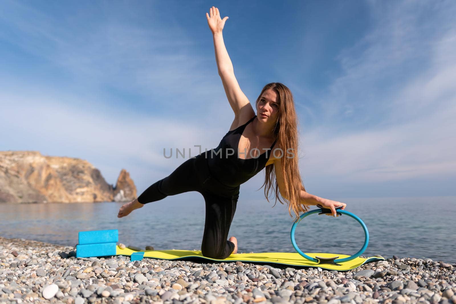 Young woman with long hair, fitness instructor in black Sportswear Leggings and Tops, stretching on a yoga mat with magic pilates ring near the sea on a sunny day, female fitness yoga routine concept.