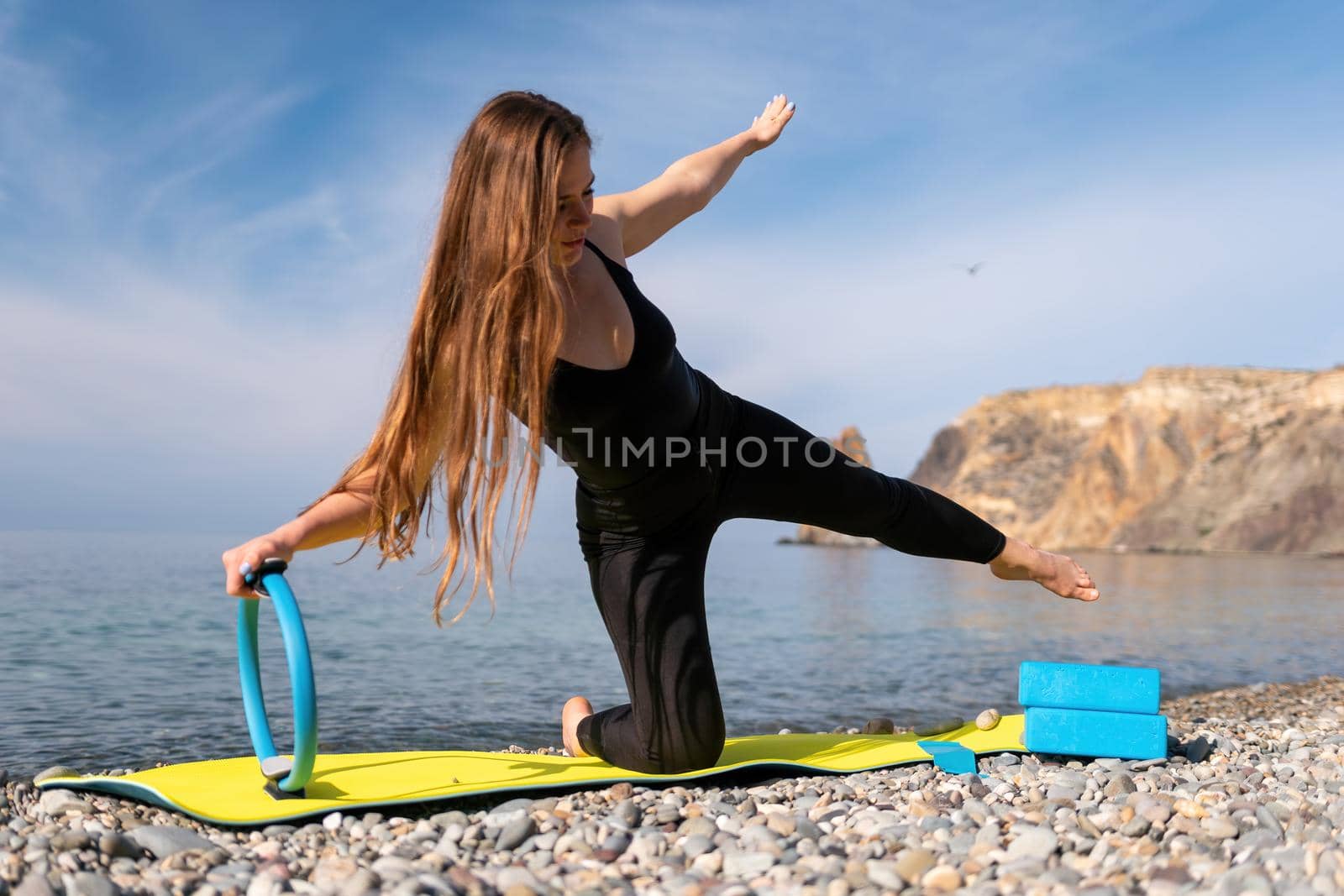 Young woman with long hair, fitness instructor in black Sportswear Leggings and Tops, stretching on a yoga mat with magic pilates ring near the sea on a sunny day, female fitness yoga routine concept.