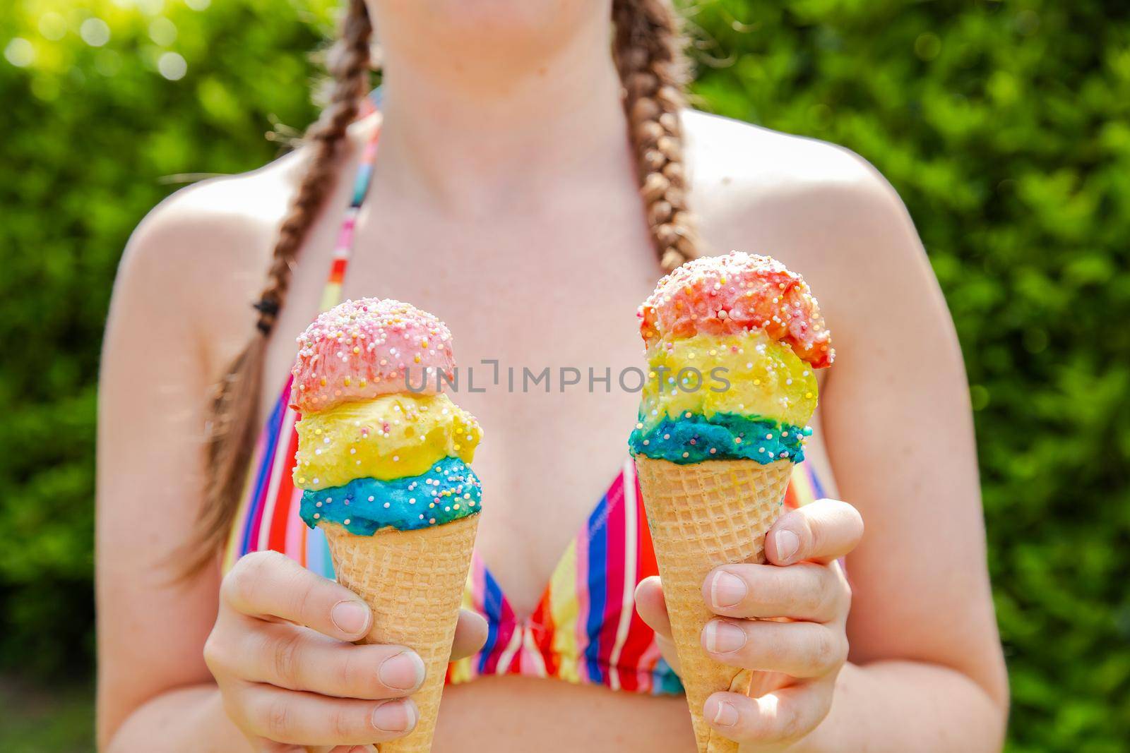 Beautiful girl holding two colorful ice cream with sprinkles wearing a rainbow colored swimsuit in the summer, pink sunglasses and braids happy vacation,holidays concept sunshine