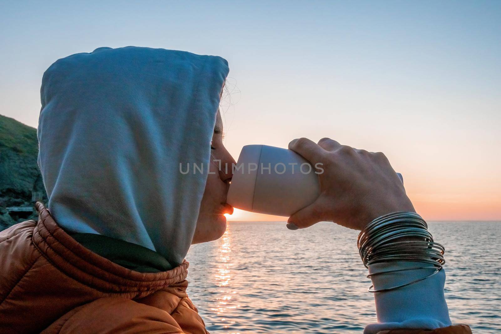 A young tourist Woman holding coffee tumbler cup while sitting outdoor and enjoying sunrise over sea mountain landscape. Women's yoga fitness routine. Healthy lifestyle, harmony and meditation by panophotograph