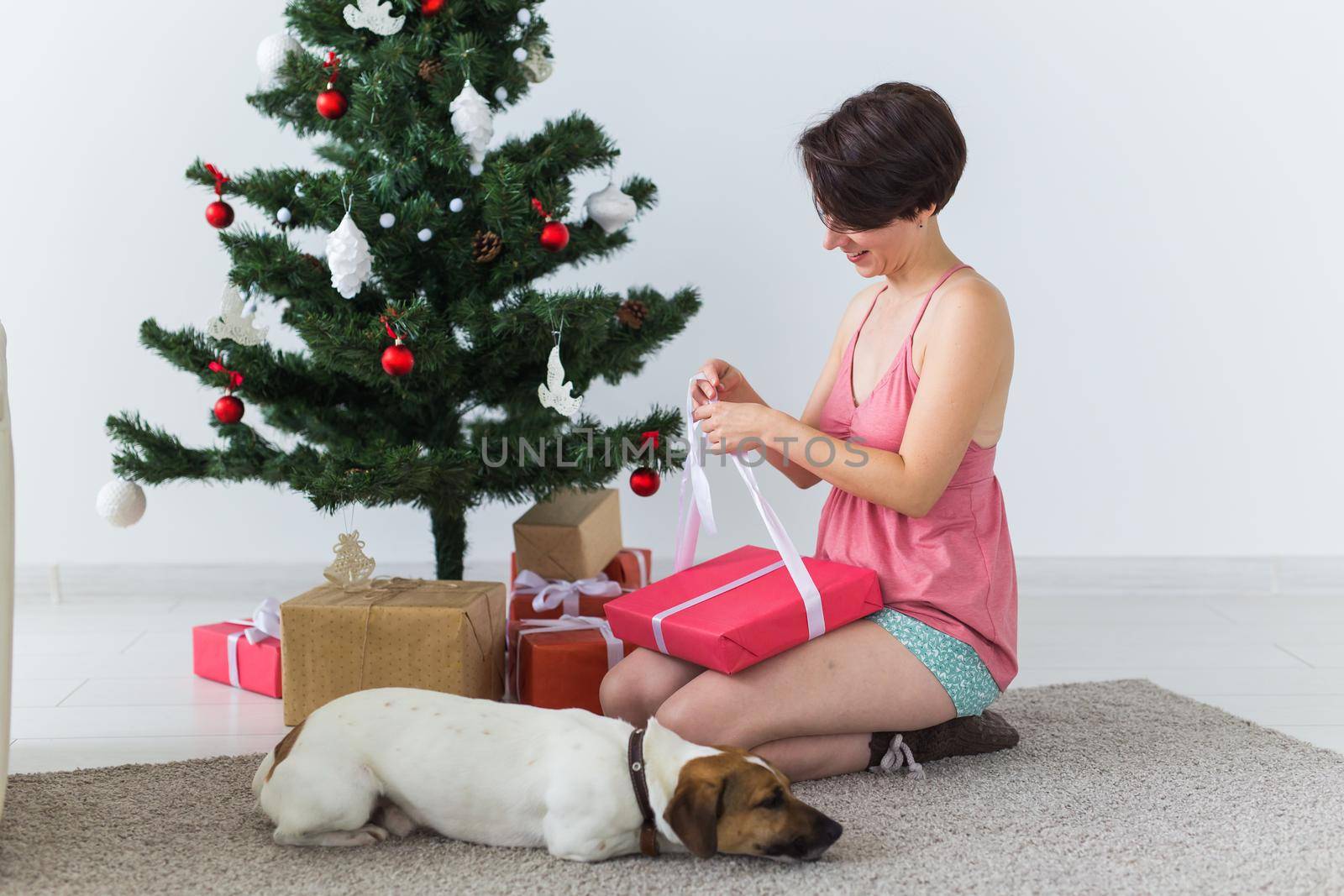 Happy woman with dog opening Christmas gifts. Christmas tree with presents under it. Decorated living room.