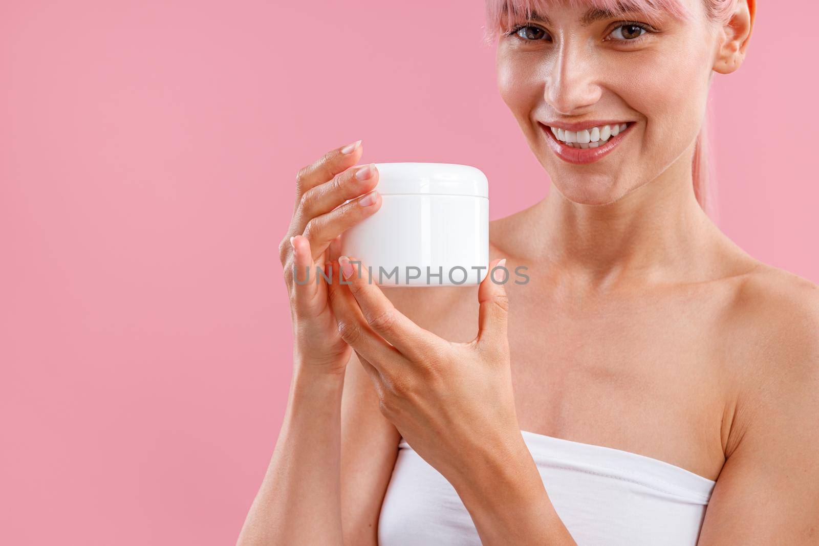 Cropped portrait of young woman smiling at camera, holding white jar with moisturizing body lotion after shower isolated over pink background. Beauty, spa, body care concept