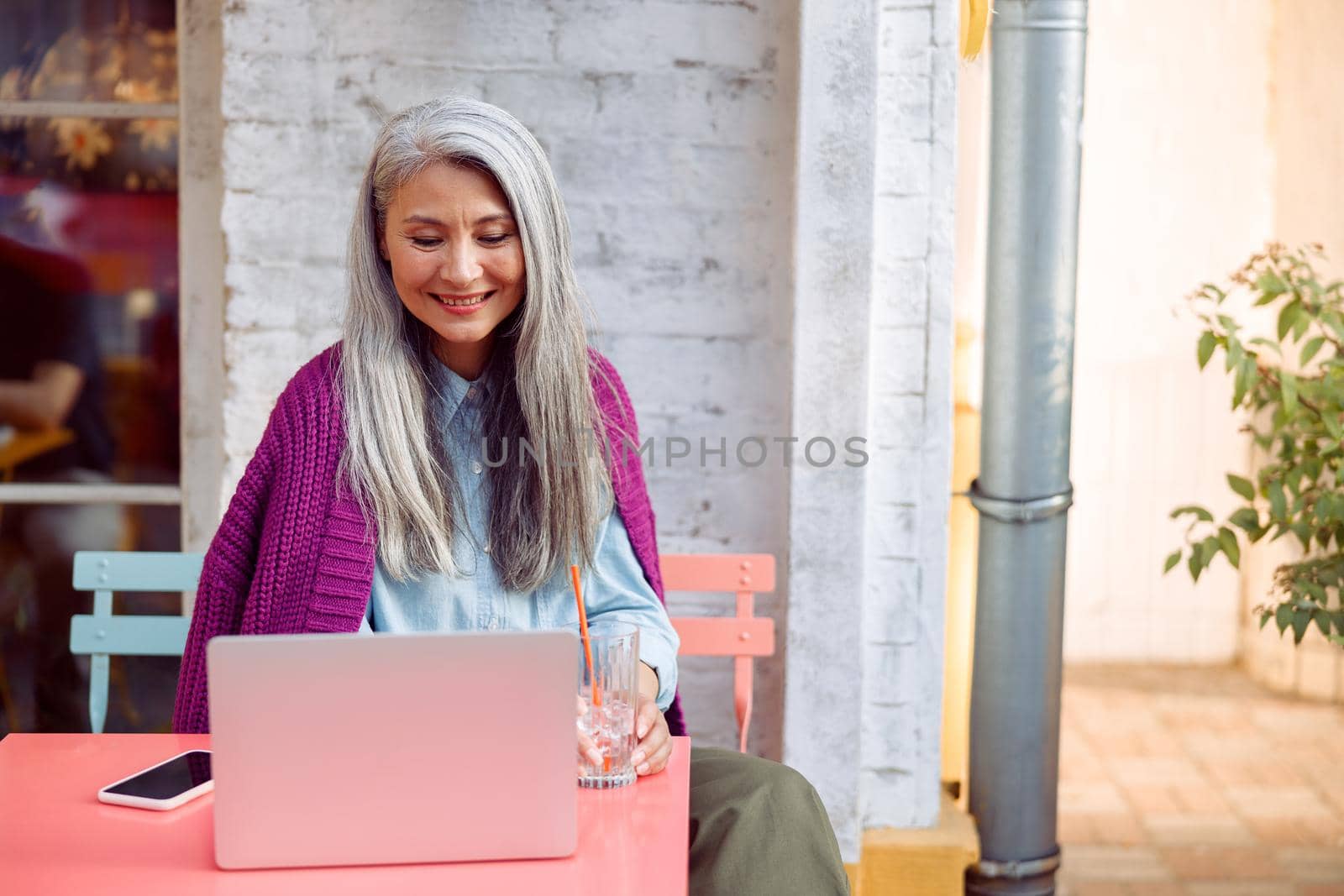 Positive senior Asian woman with glass of water and smartphone looks at laptop screen at coral table on outdoors cafe terrace