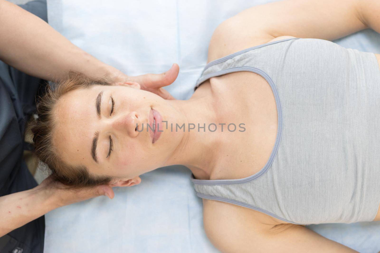 Young woman with closed eyes having a chiropractic treatment in the clinic - lying on the couch and the doctor holding her neck. Mid shot