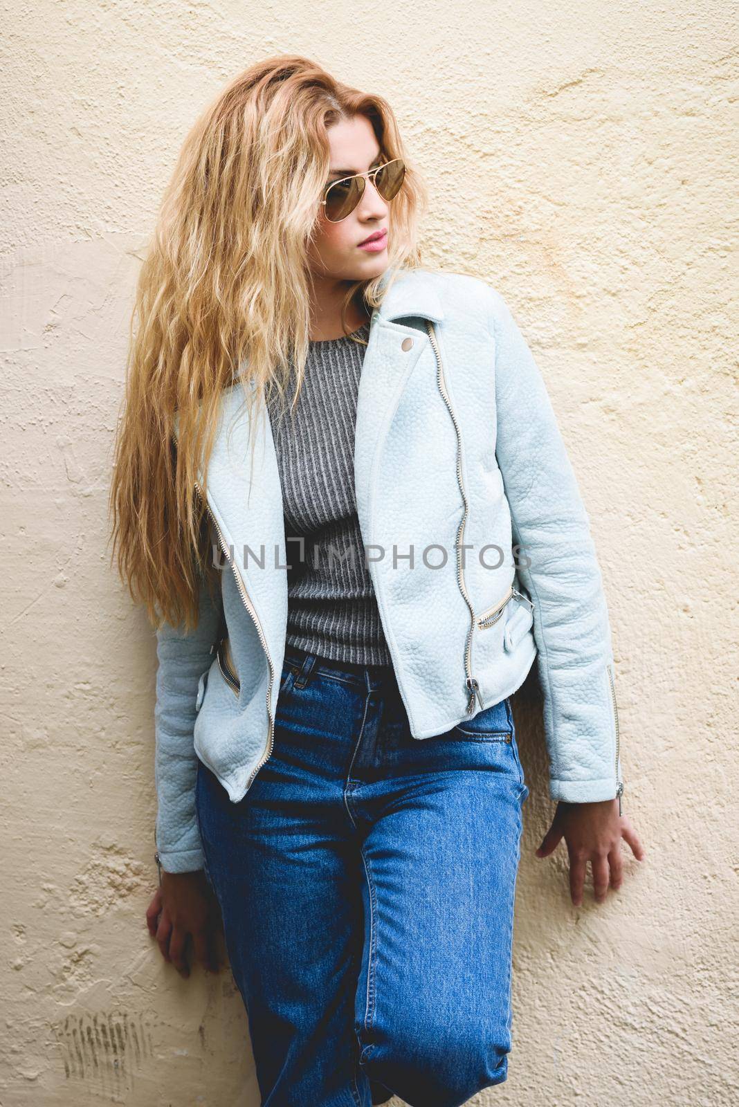 Portrait of beautiful young blonde woman with curly hair. Girl wearing leather jacket, blue jeans and aviator sunglasses in urban background.