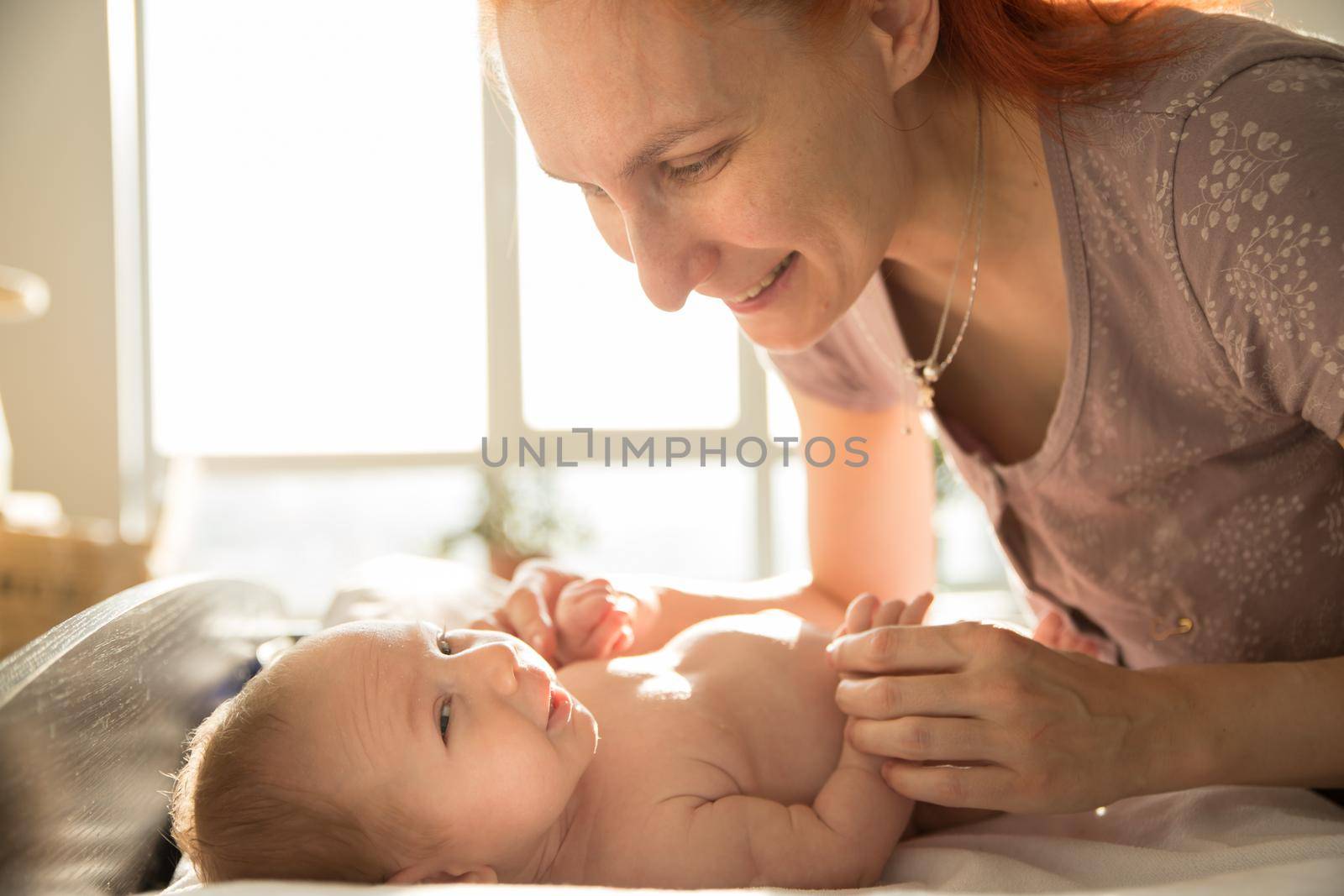 Smiling ginger woman mother takes care of her small baby lying on a bed. Mid shot