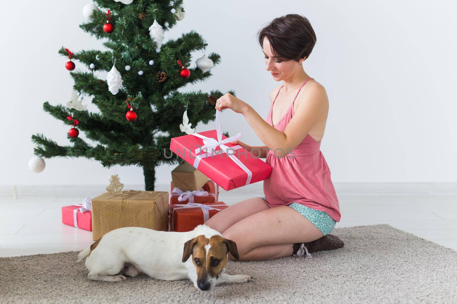 Happy young woman with lovely dog opening present box under christmas tree.