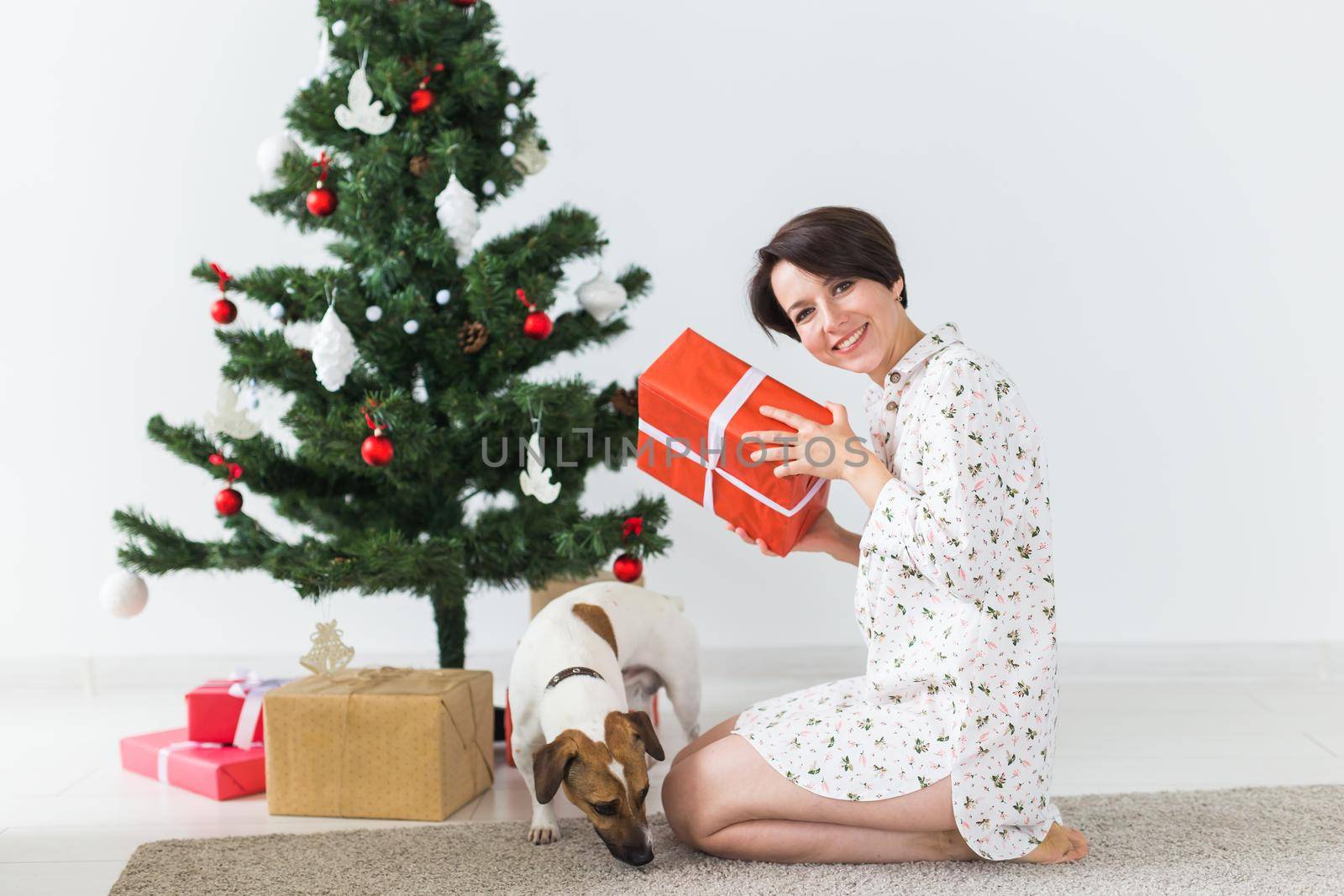 Happy woman with dog opening Christmas gifts. Christmas tree with presents under it. Decorated living room by Satura86