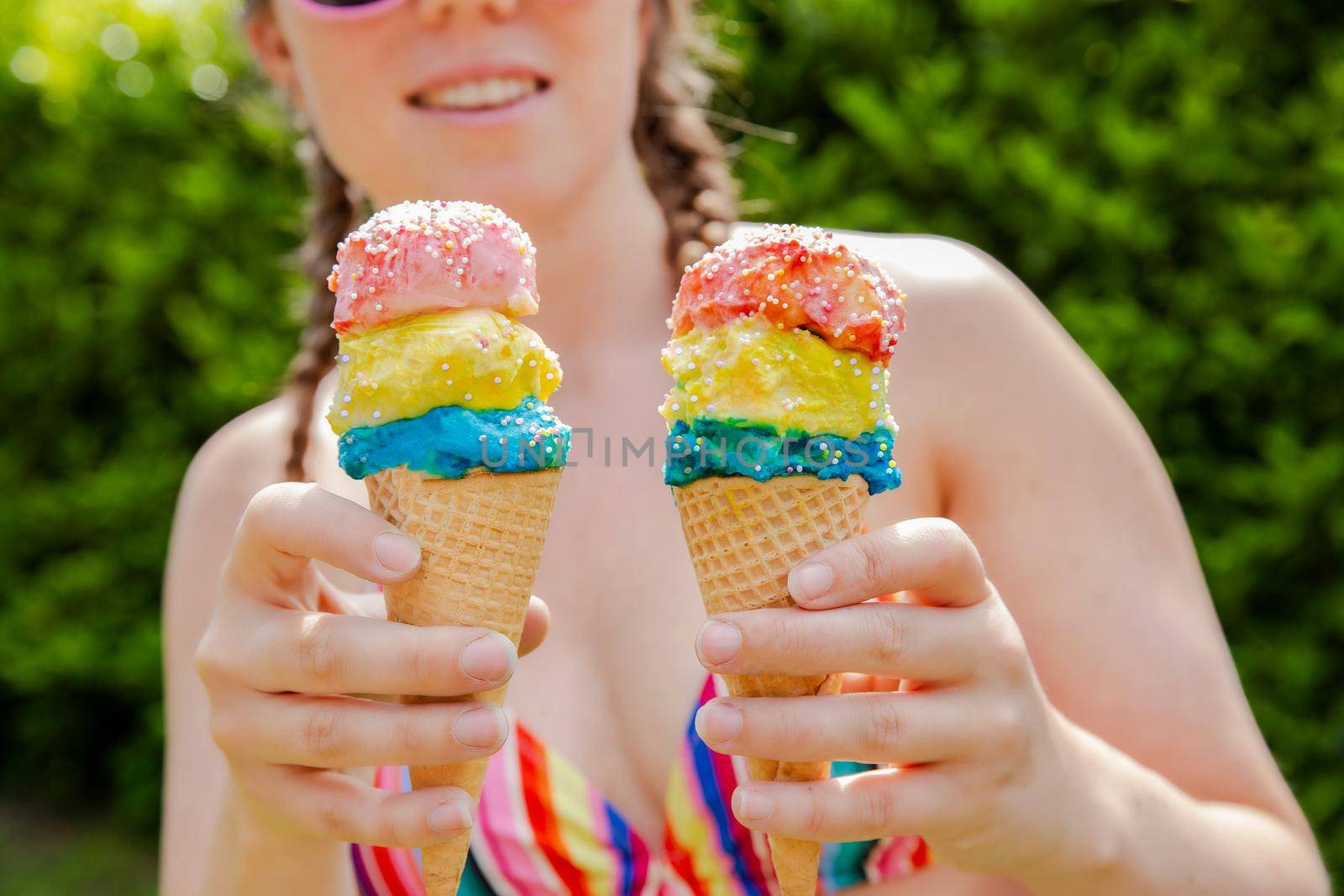 Beautiful girl holding two colorful ice cream with sprinkles wearing a rainbow colored swimsuit in the summer, pink sunglasses and braids happy vacation,holidays concept sunshine