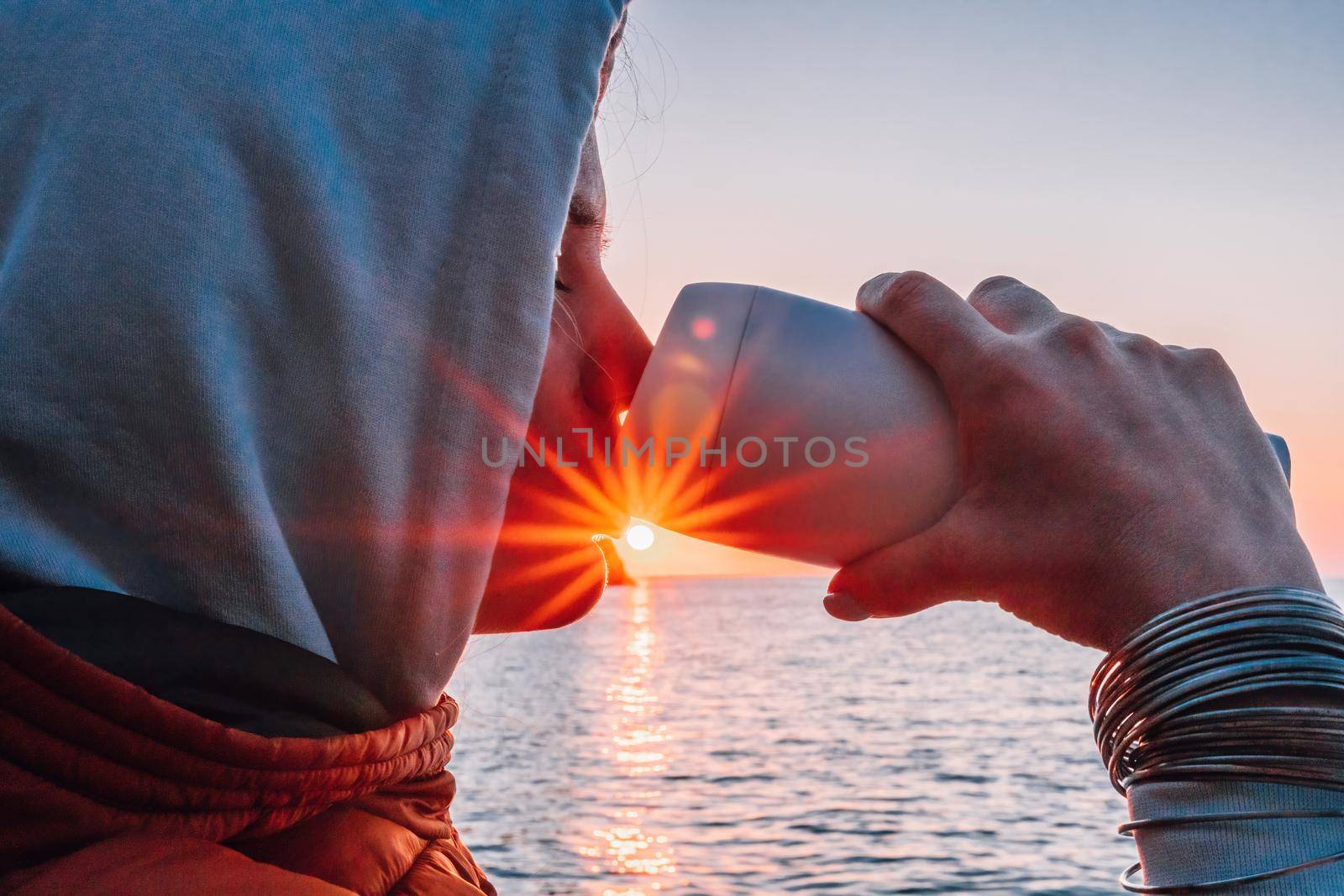 A young tourist Woman holding coffee tumbler cup while sitting outdoor and enjoying sunrise over sea mountain landscape. Women's yoga fitness routine. Healthy lifestyle, harmony and meditation by panophotograph