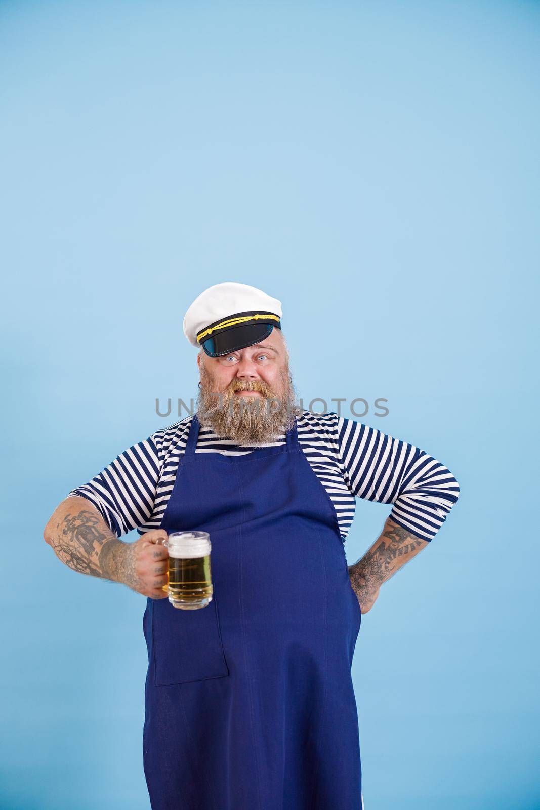 Confident man with overweight in sailor suit holds mug of tasty beer on light blue background by Yaroslav_astakhov