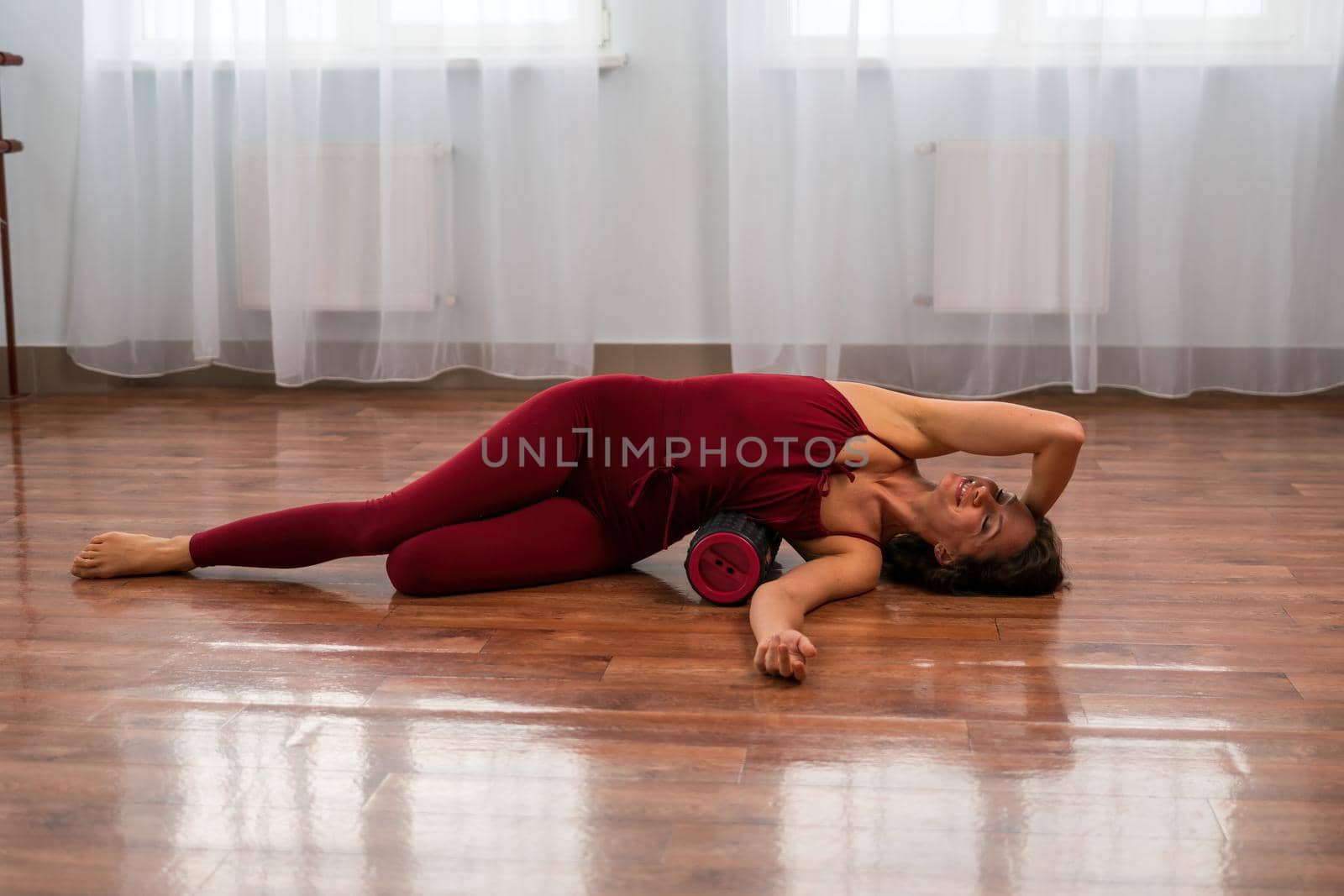 A young woman fitness instructor in red Sportswear Leggings and Top stretching in the gym before her pilates, on a yoga mat near the large window on a sunny day, female fitness yoga routine concept