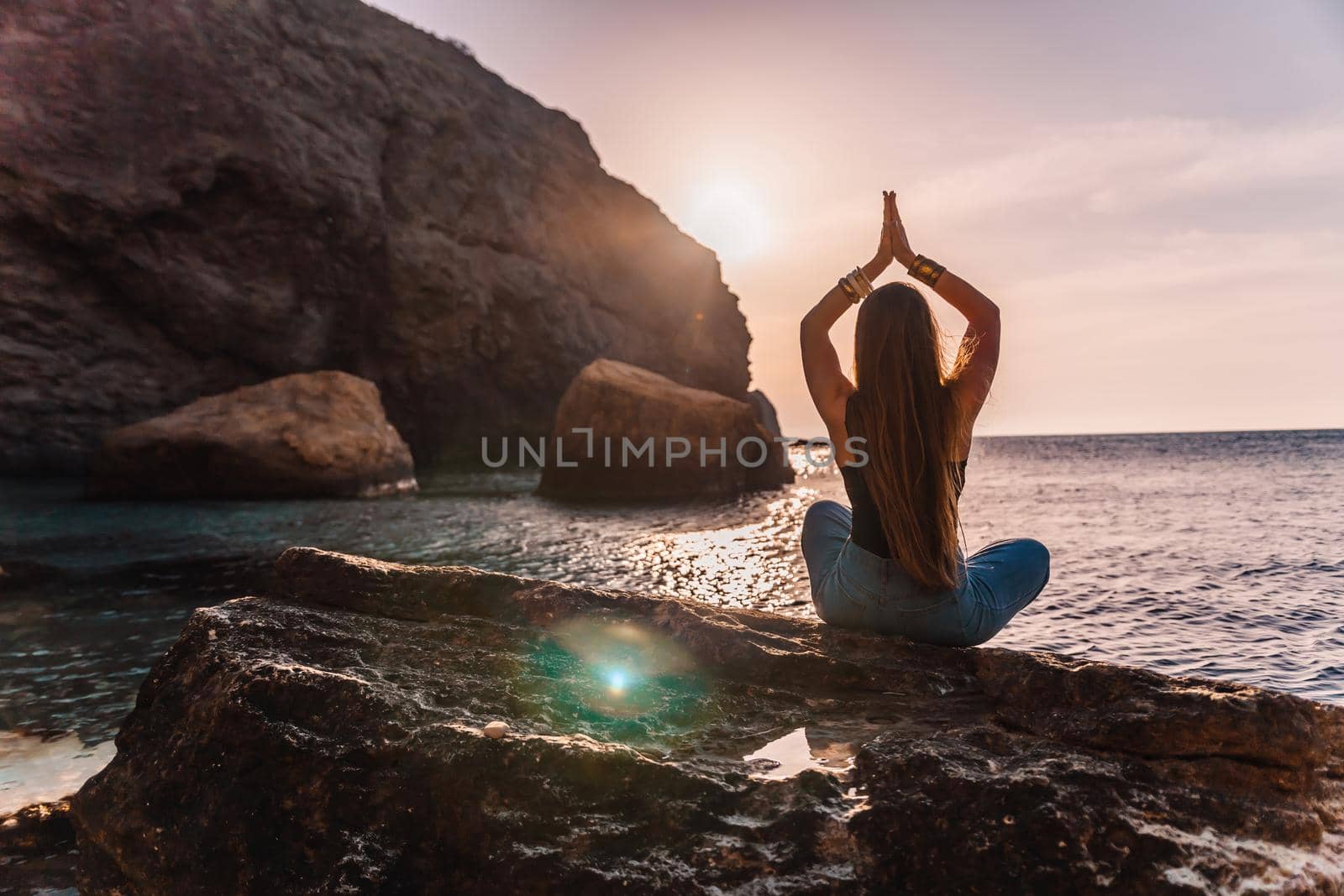 Young woman with long hair in black swimsuit and boho style braclets practicing outdoors on yoga mat by the sea on a sunset. Women's yoga fitness routine. Healthy lifestyle, harmony and meditation by panophotograph