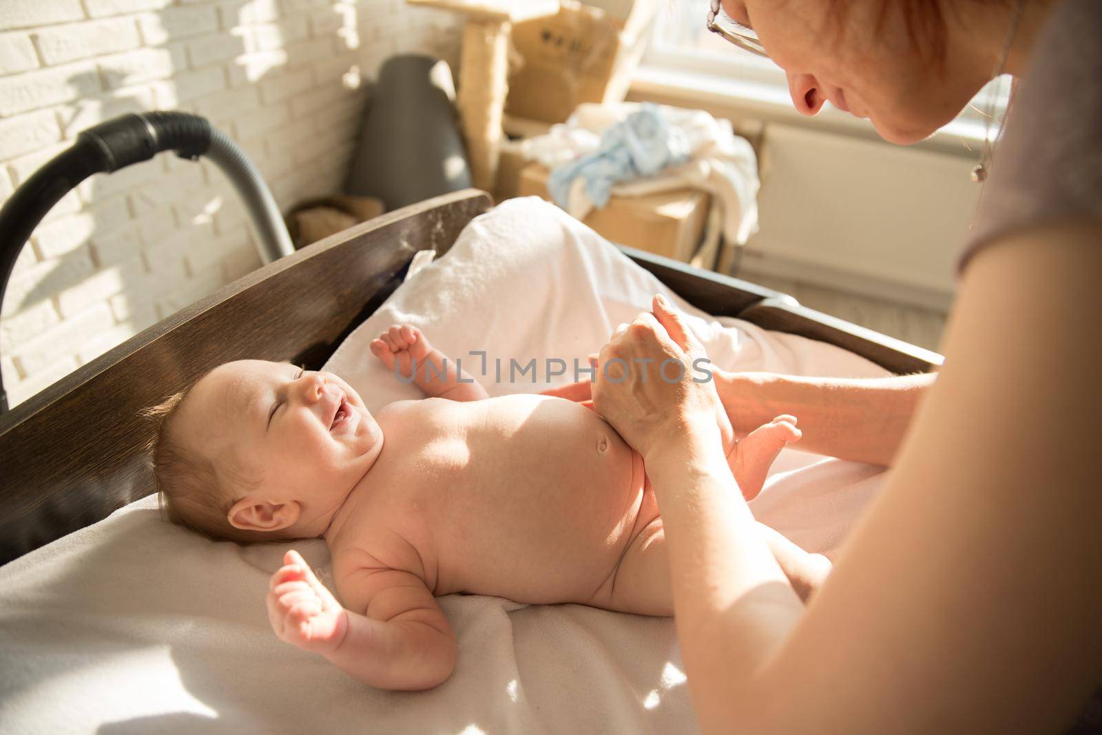 Mother massaging legs of her smiling baby lying on a bed. Mid shot
