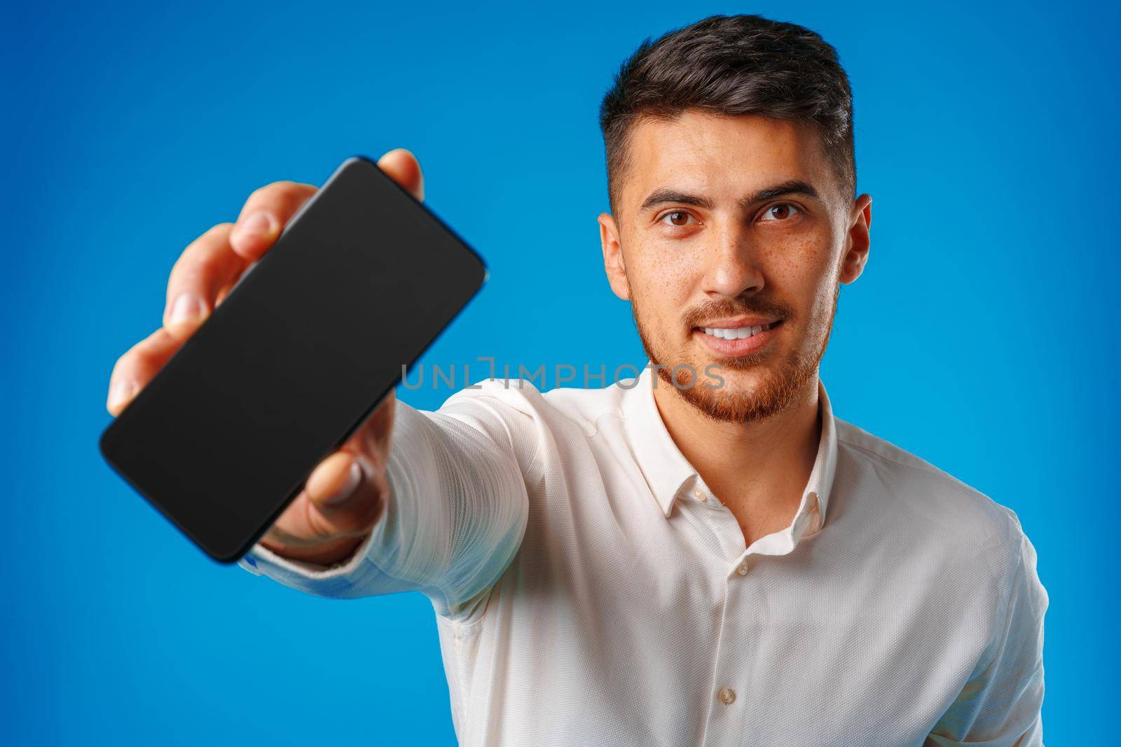 Young handsome businessman showing black smartphone screen against blue background close up