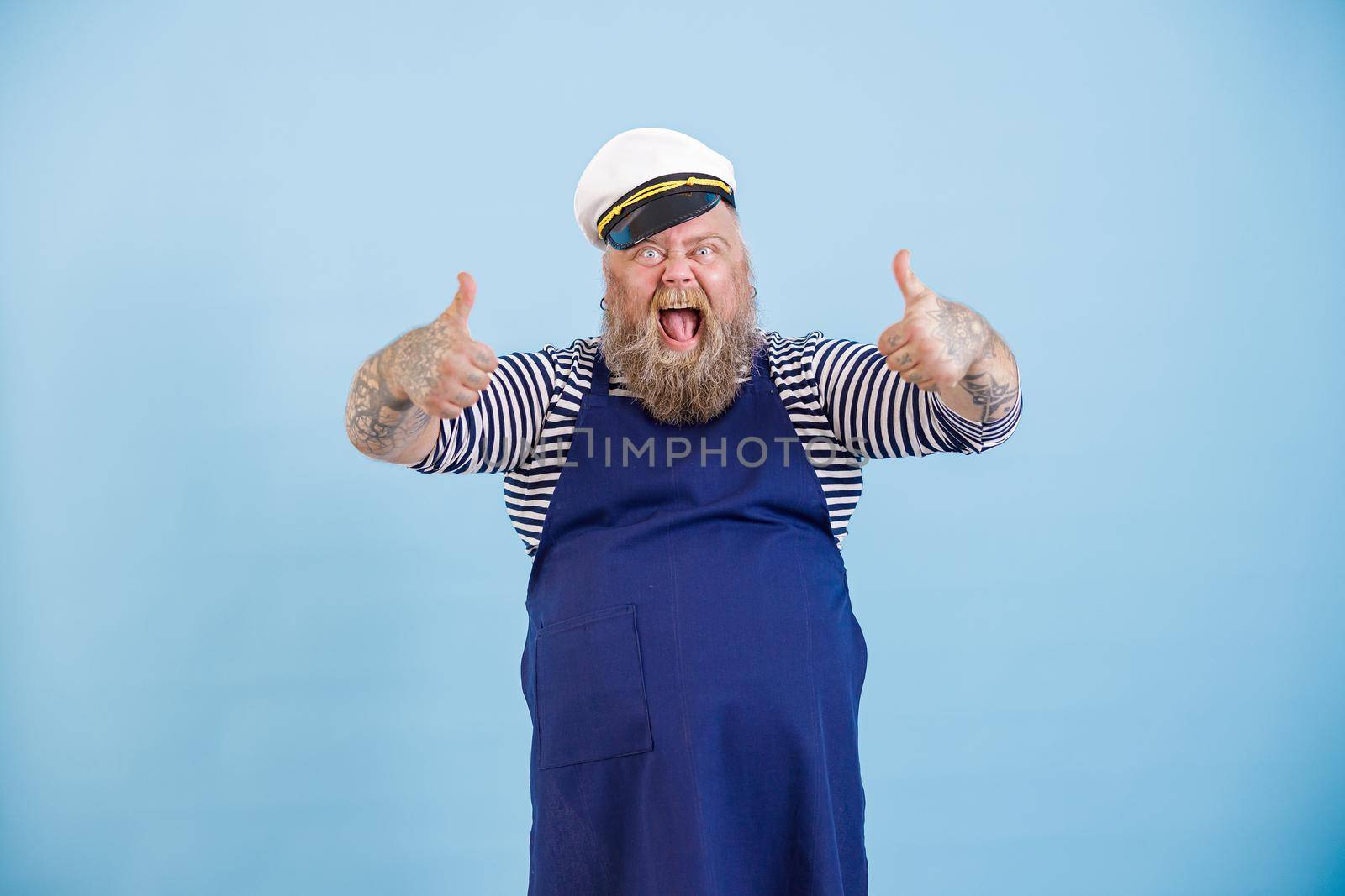 Screaming emotional male person with overweight in sailor suit and apron shows thumb up on light blue background in studio