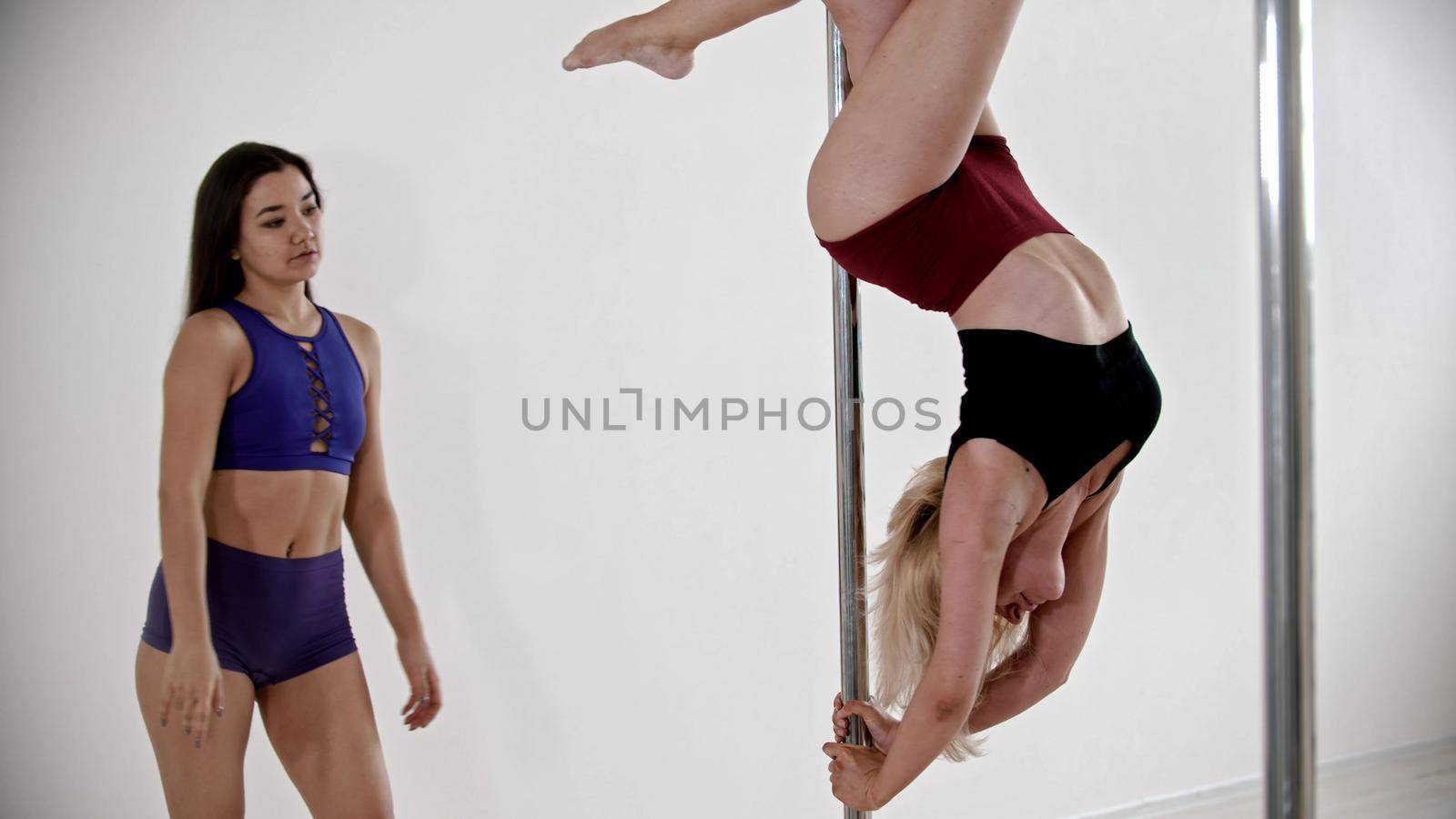 A blonde woman having a pole dancing training in the studio - keeping the balance upside down - her trainer watching her. Mid shot