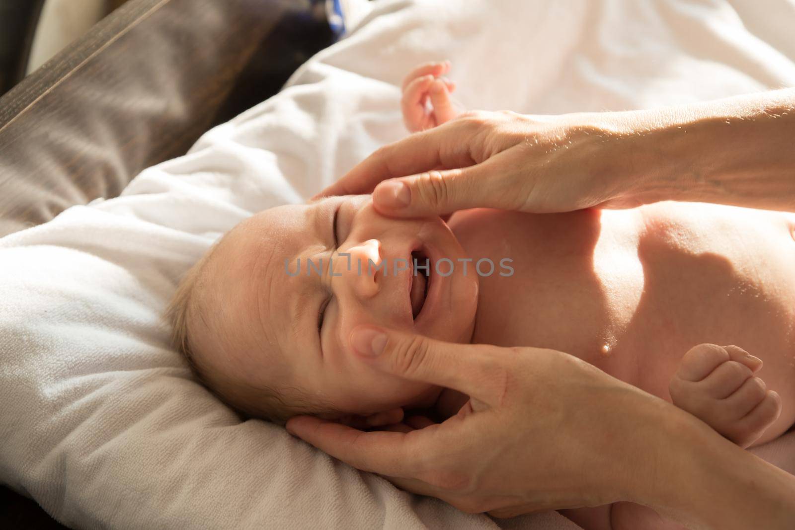 A little crying baby lying on a bed on white sheets while his parent massaging his face. Mid shot
