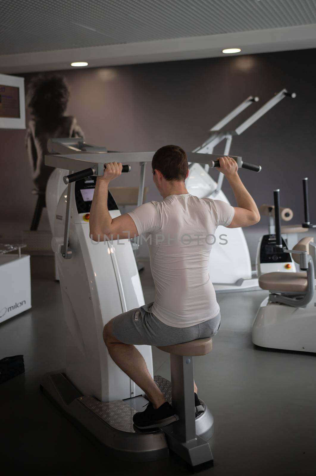 Young man in white t-shirt sitting on the strength training machine and pumping his hands muscles. Mid shot