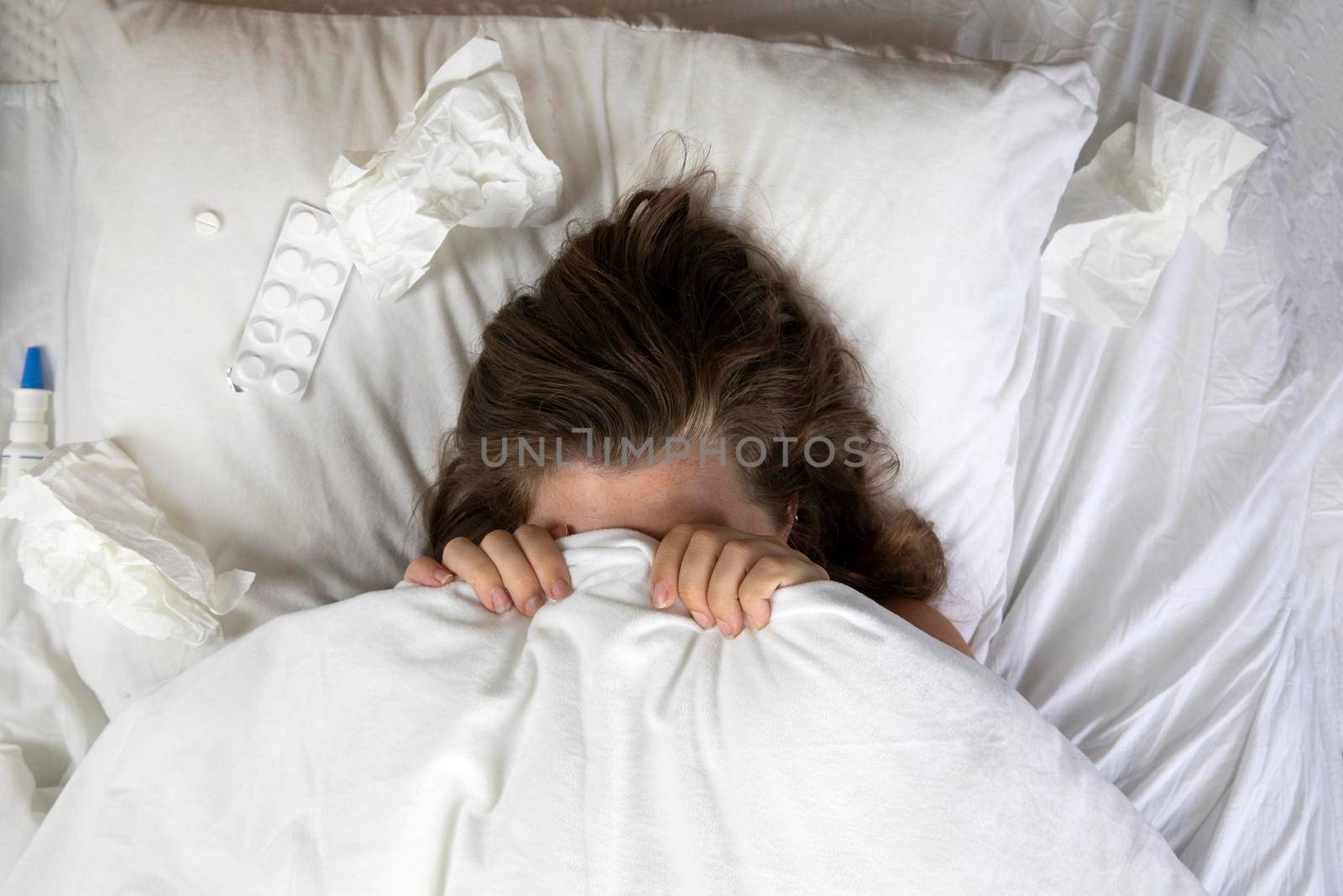 A sick young woman in bed, lying with her head under the blankets and a pile of tissues next to her top view. Fever staying home from work. white sheets and pillow