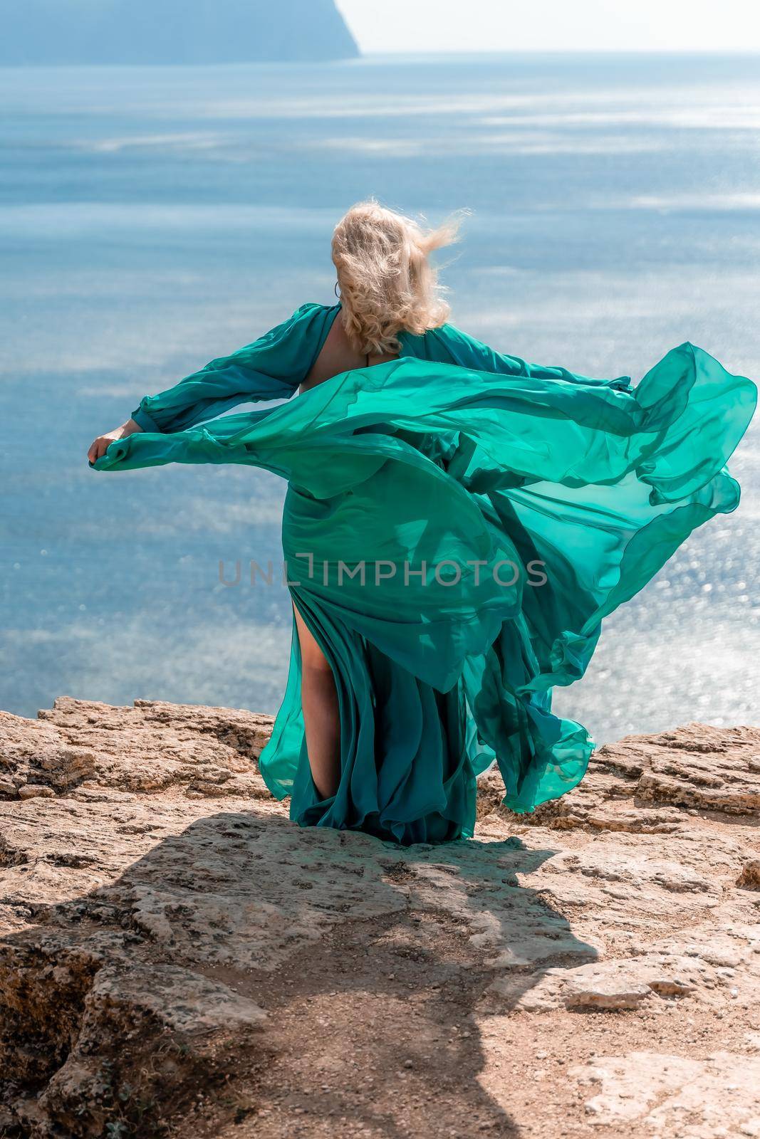 A beautiful young woman in a mint light dress with long legs stands on the edge of a cliff above the sea waving long dress, against the background of the blue sky and the sea. by Matiunina