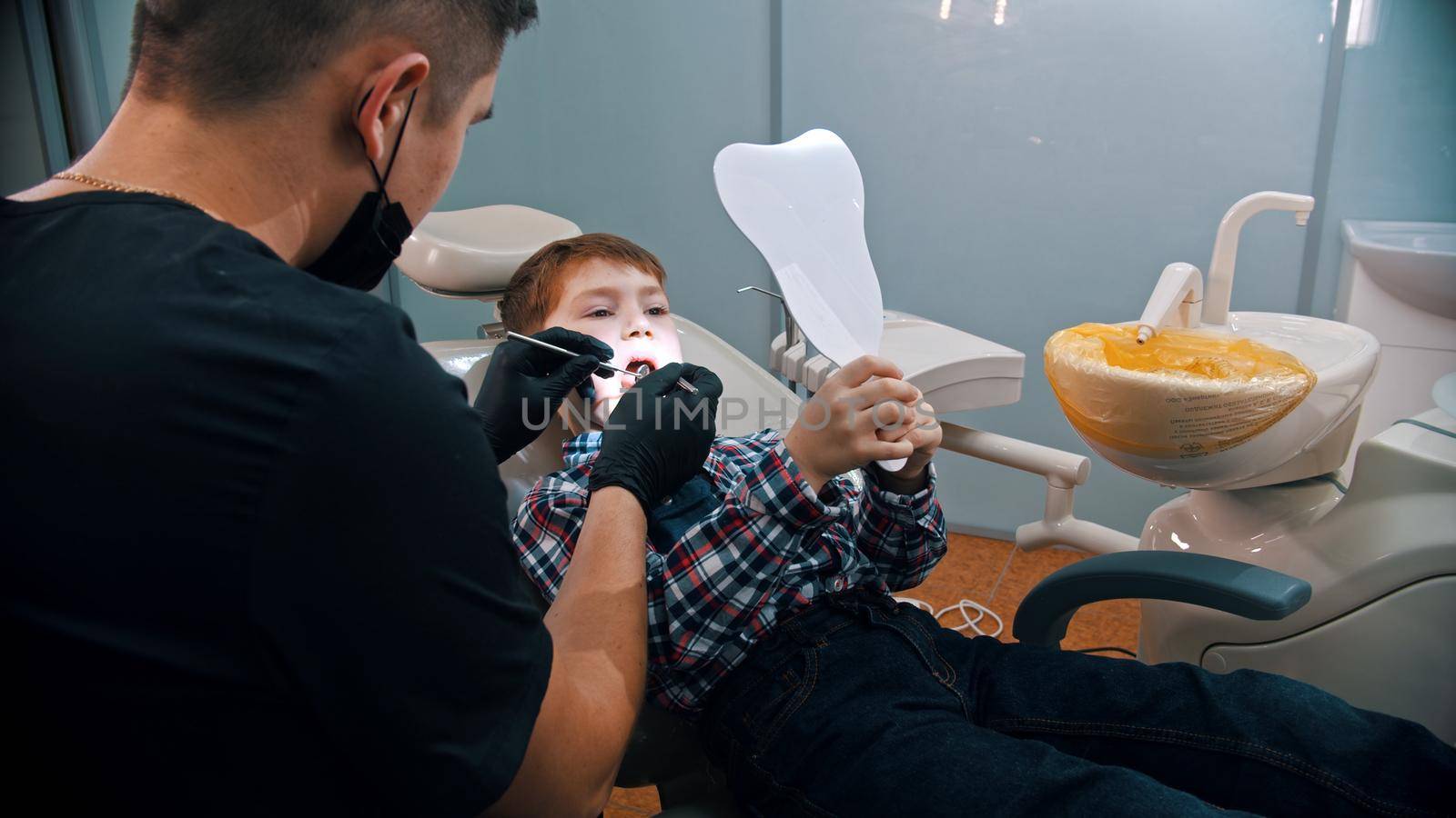 A little boy having a treatment in the modern dentistry - the boy holding a mirror and looking at his reflection. Mid shot