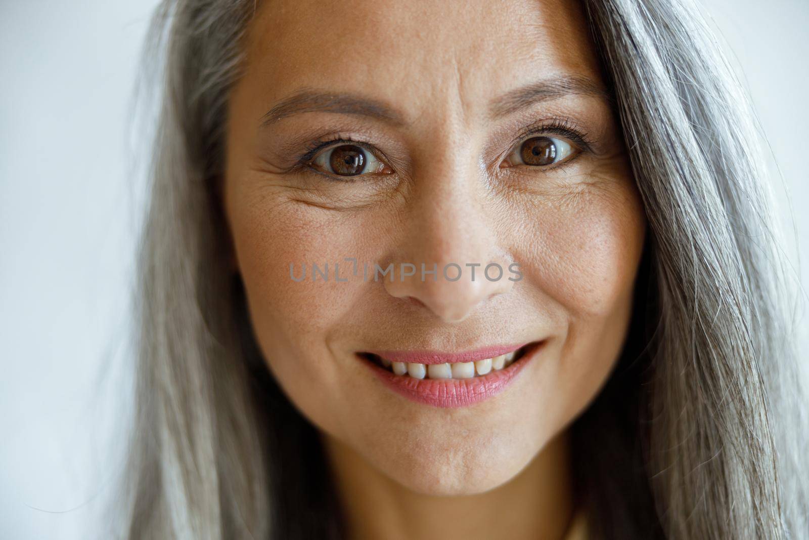 Cheerful silver haired lady with light makeup looks into camera on light grey background in studio closeup. Mature beauty lifestyle