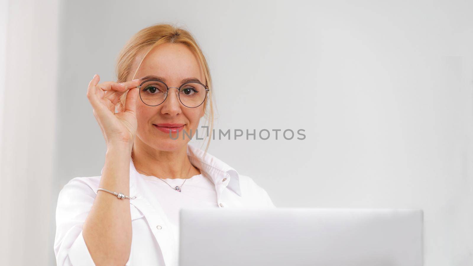 A blonde doctor in glasses or a cosmetologist in a white coat is sitting at a computer. She reads and types on the computer. Concept of medicine, beauty, cosmetologist, masseur. by Matiunina