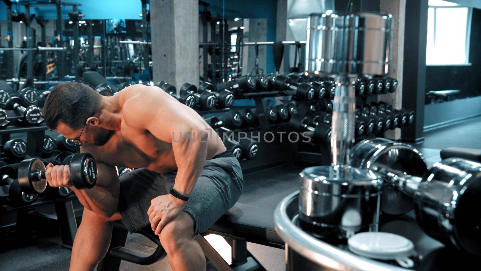 A man bodybuilder pumping his hand muscles sitting in the sports gym. Mid shot