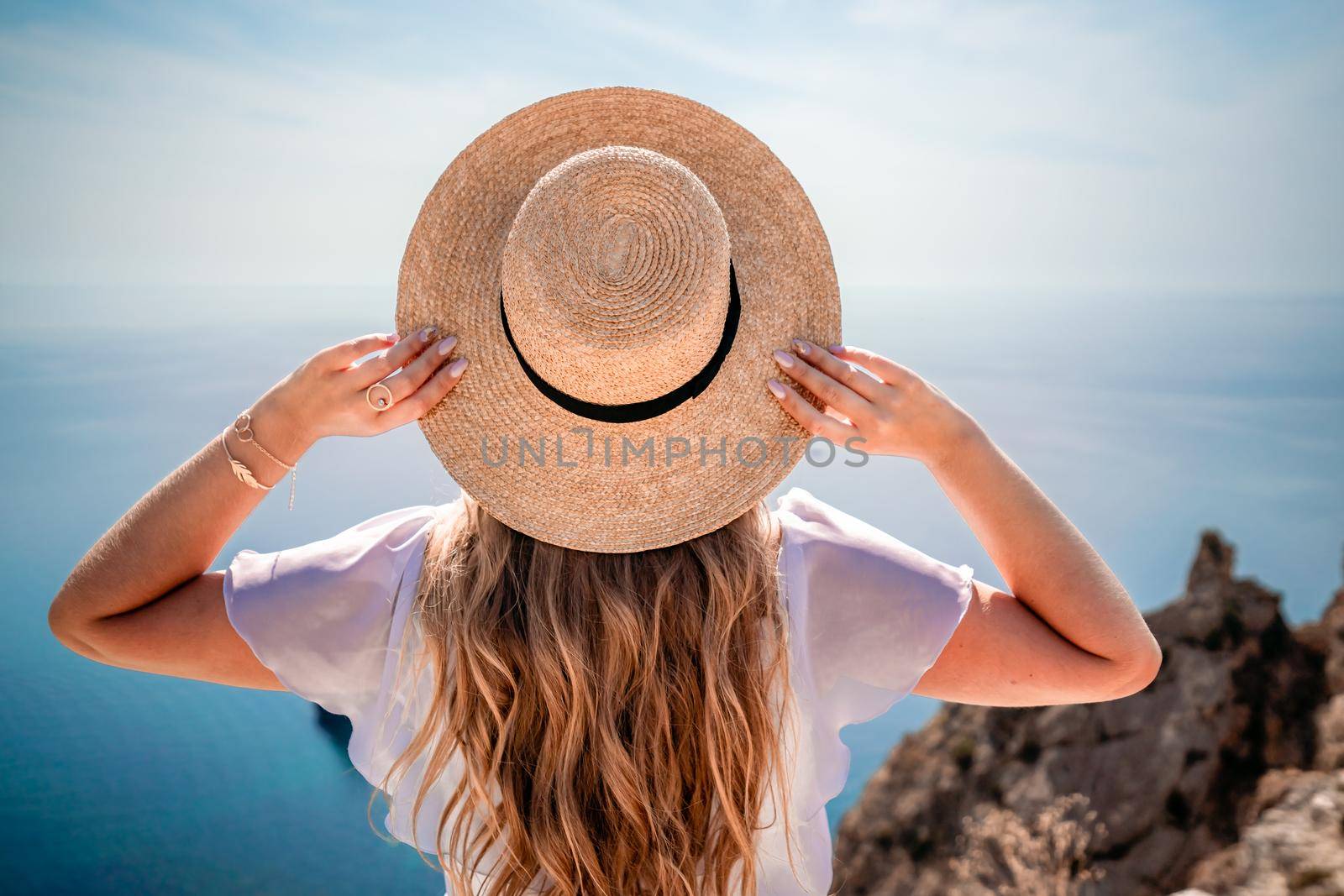 Woman enjoying summer beach vacation on the background of the ocean. A woman adjusts her straw hat on her head while sitting facing the sea