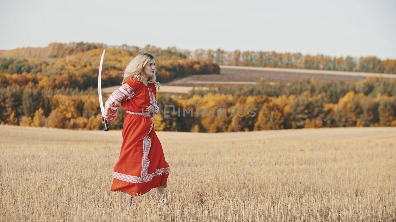 Militant woman in red clothes stands in a field with a sword. Mid shot
