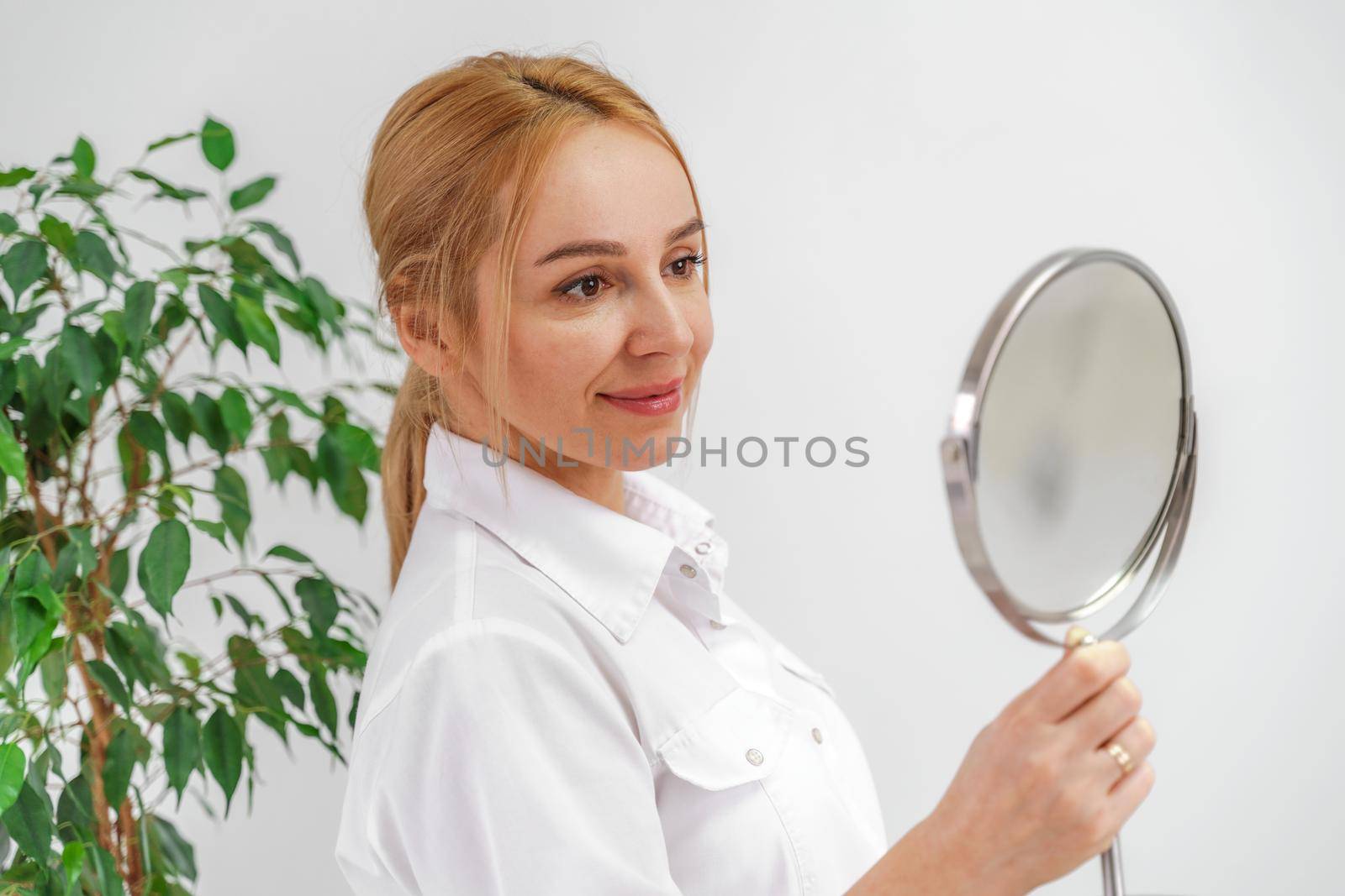 A blonde woman in white formal clothes looks at herself in the mirror. On a white background, green leaves at the back