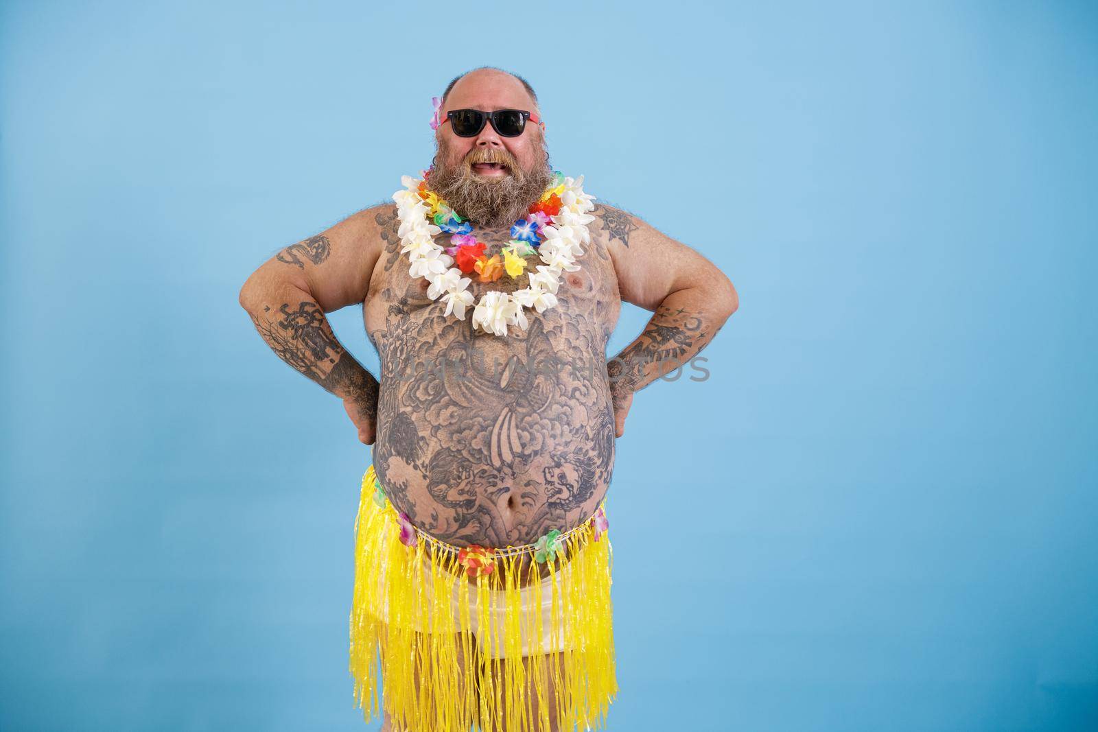 Happy bearded man with overweight and tattoos on large tummy in decorative yellow grass skirt and flowers garland on light blue background in studio