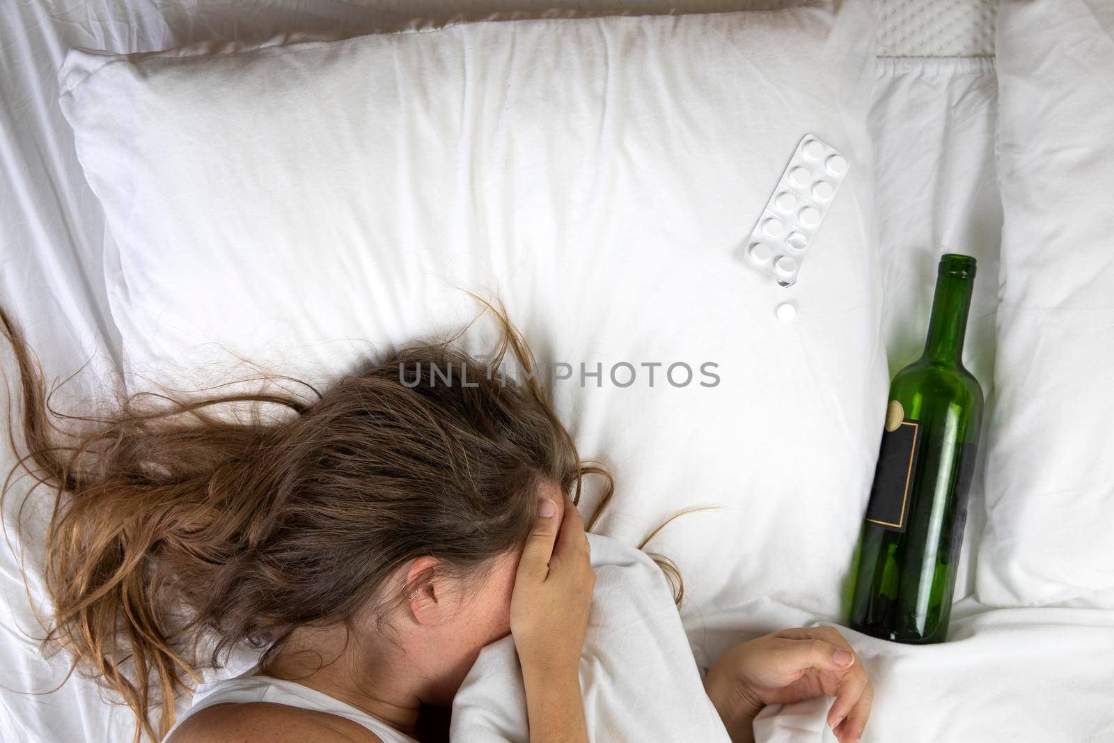 Sleeping drunk girl with empty wine bottle. Lying on the bed fainted, alcohol abuse,addiction concept top view white sheets