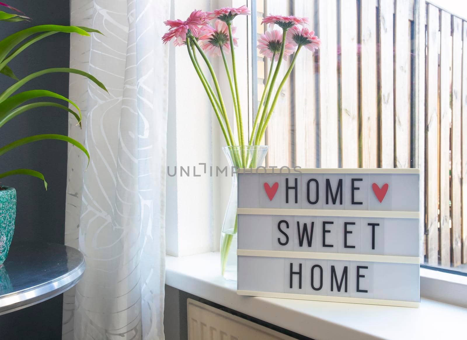 Sign Home sweet home text on lightbox ,windowsill near window with pink flowers, decorative frame modern interior background, stylish house cozy corner in lovely living room summer