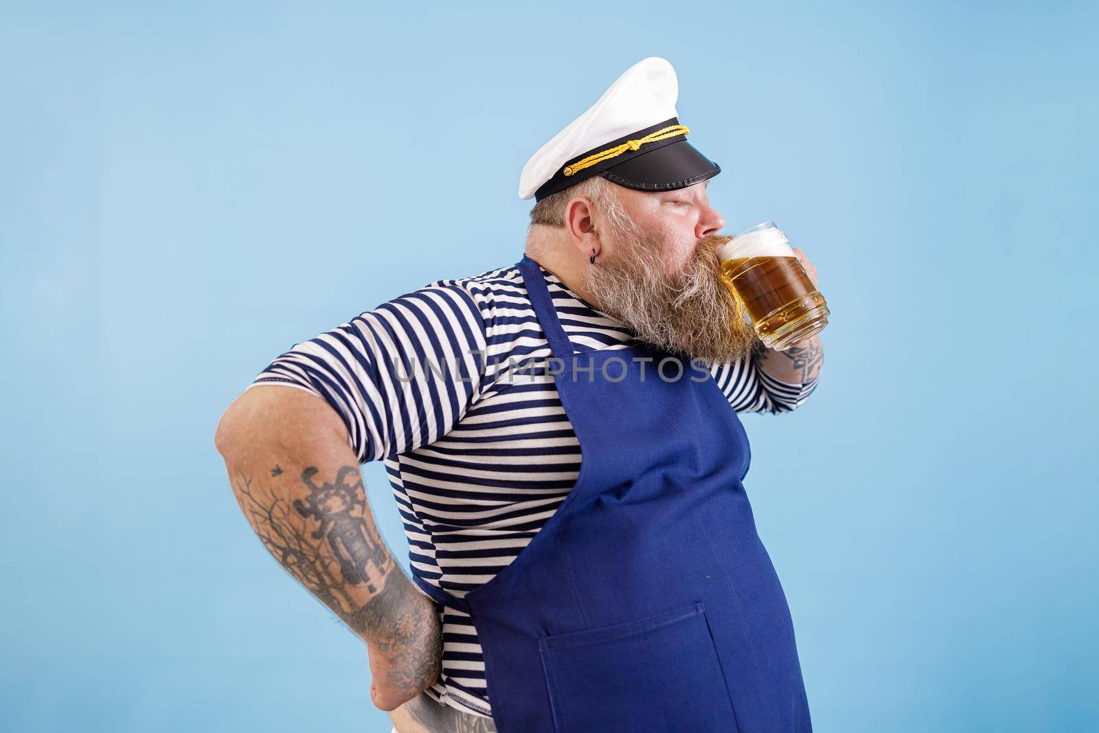 Happy mature plus size man in sailor suit with apron and captainhat drinks fresh beer on light blue background in studio side view